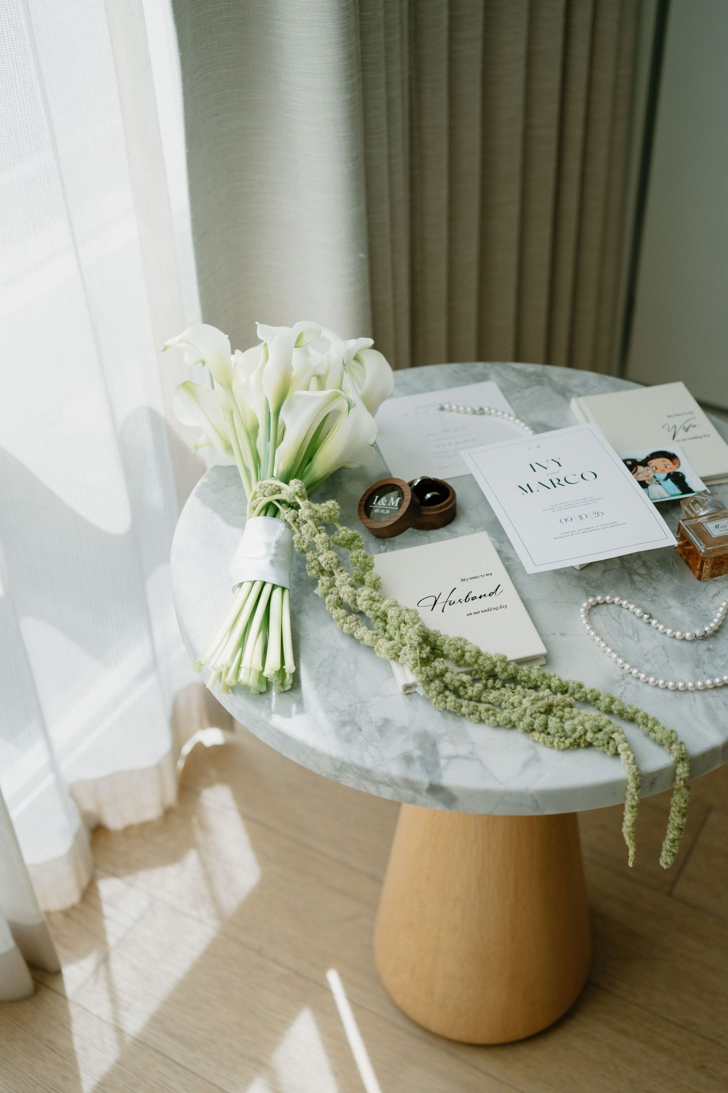 Elegant wedding flat lay with white calla lily bouquet, invitation suite, pearl necklace, and ring box on marble table.