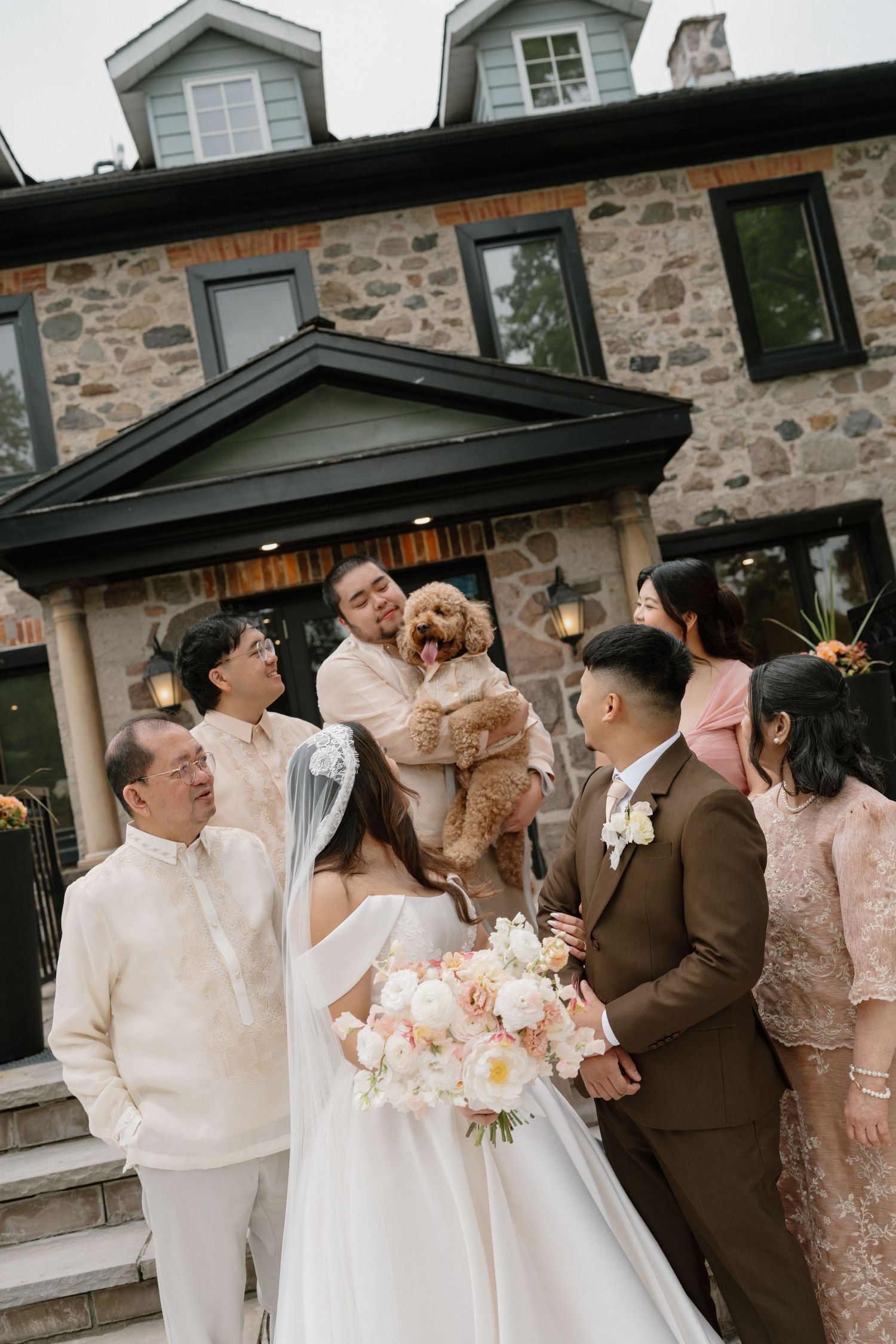 Wedding party with bride holding bouquet, groom lifting a dog, and family gathered outside a stone mansion.
