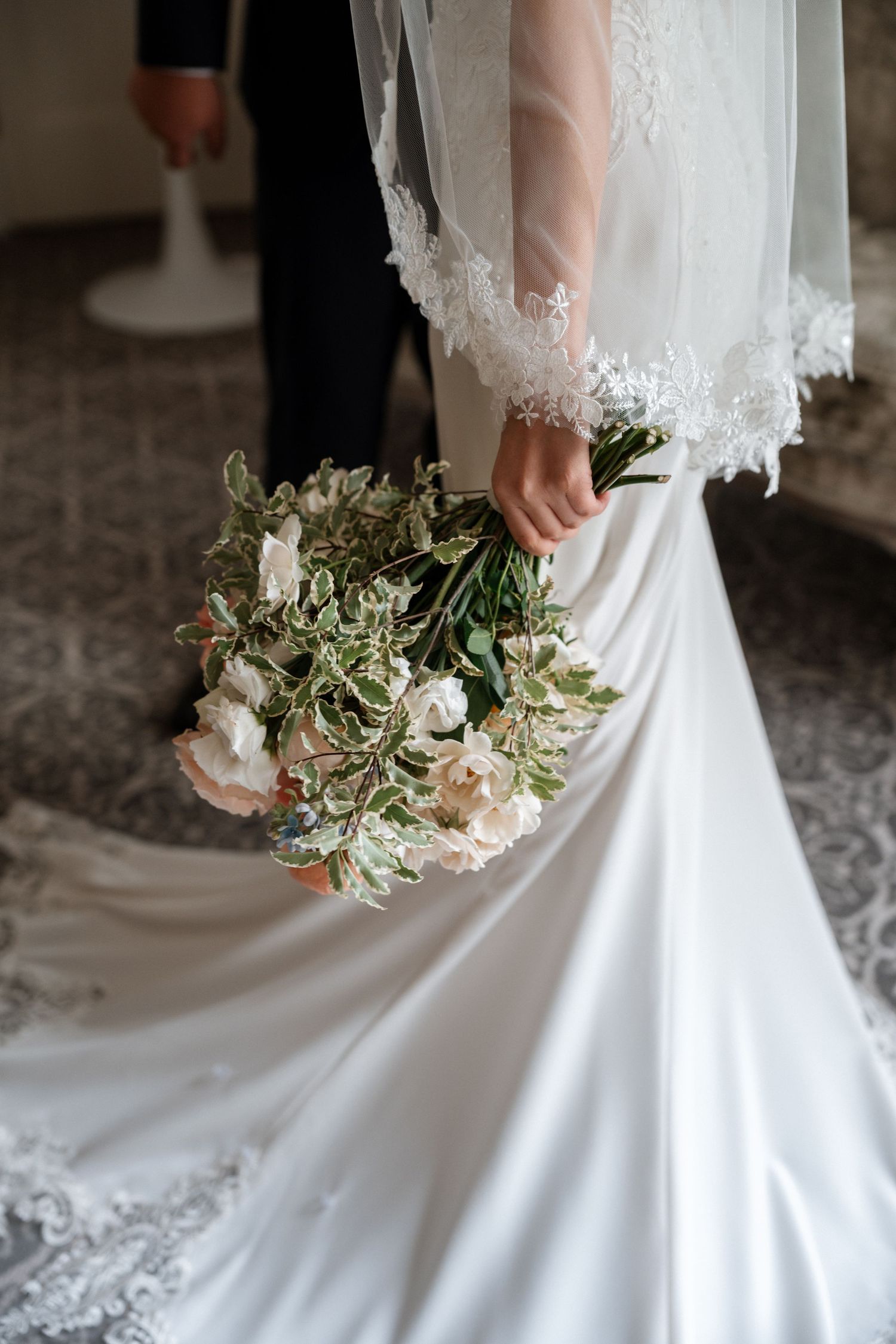 Close-up view of bride holding elegant white and green floral bouquet with trailing lace details on wedding dress.