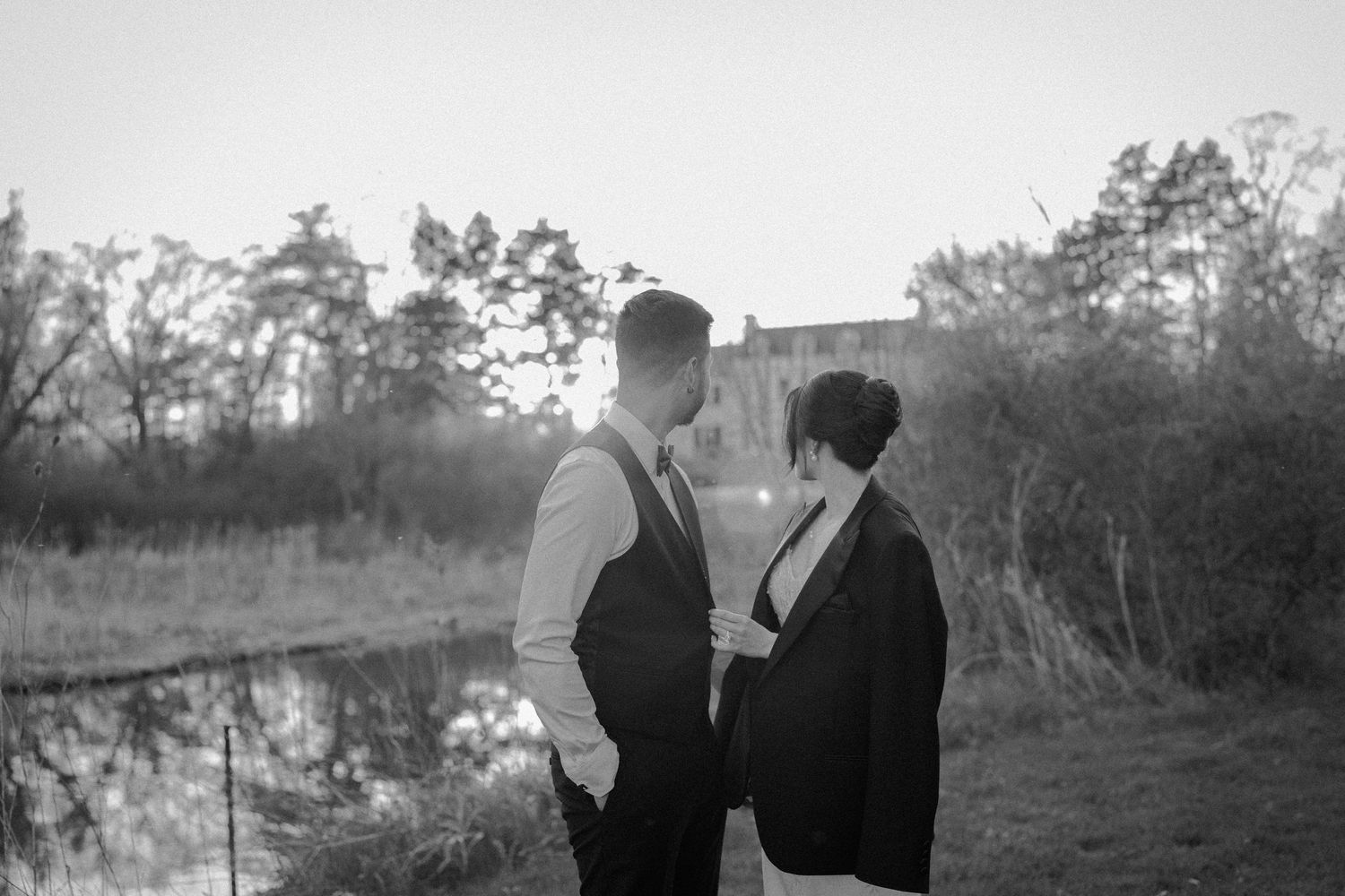 A romantic black and white portrait of a couple standing together by a lake at dusk with trees in the background.