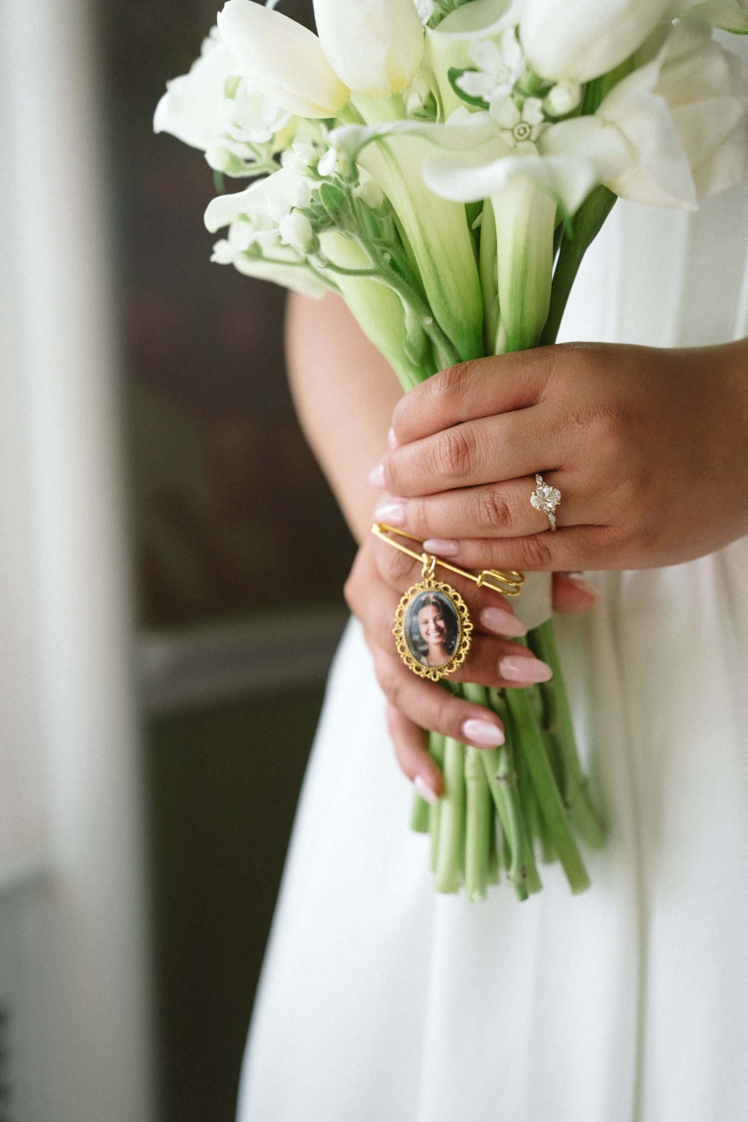 Bride holding white tulip bouquet with gold locket charm and diamond engagement ring on wedding day.