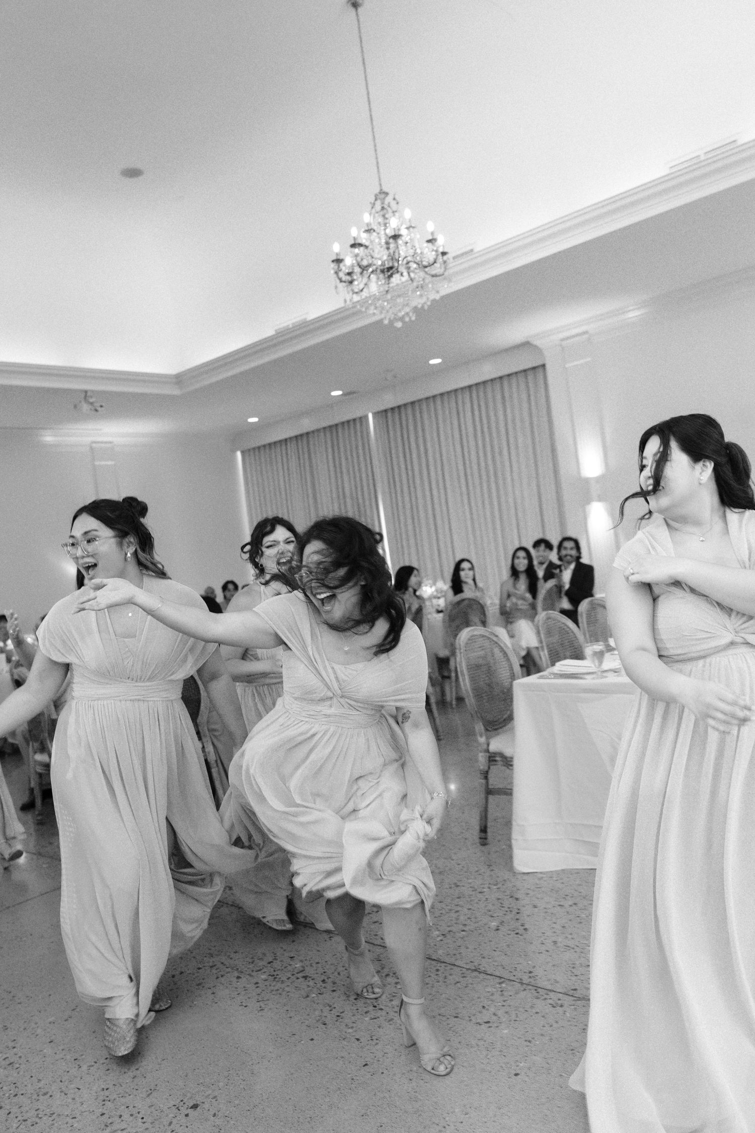 Bridesmaids in light dresses dancing energetically at a wedding reception in an elegant ballroom.