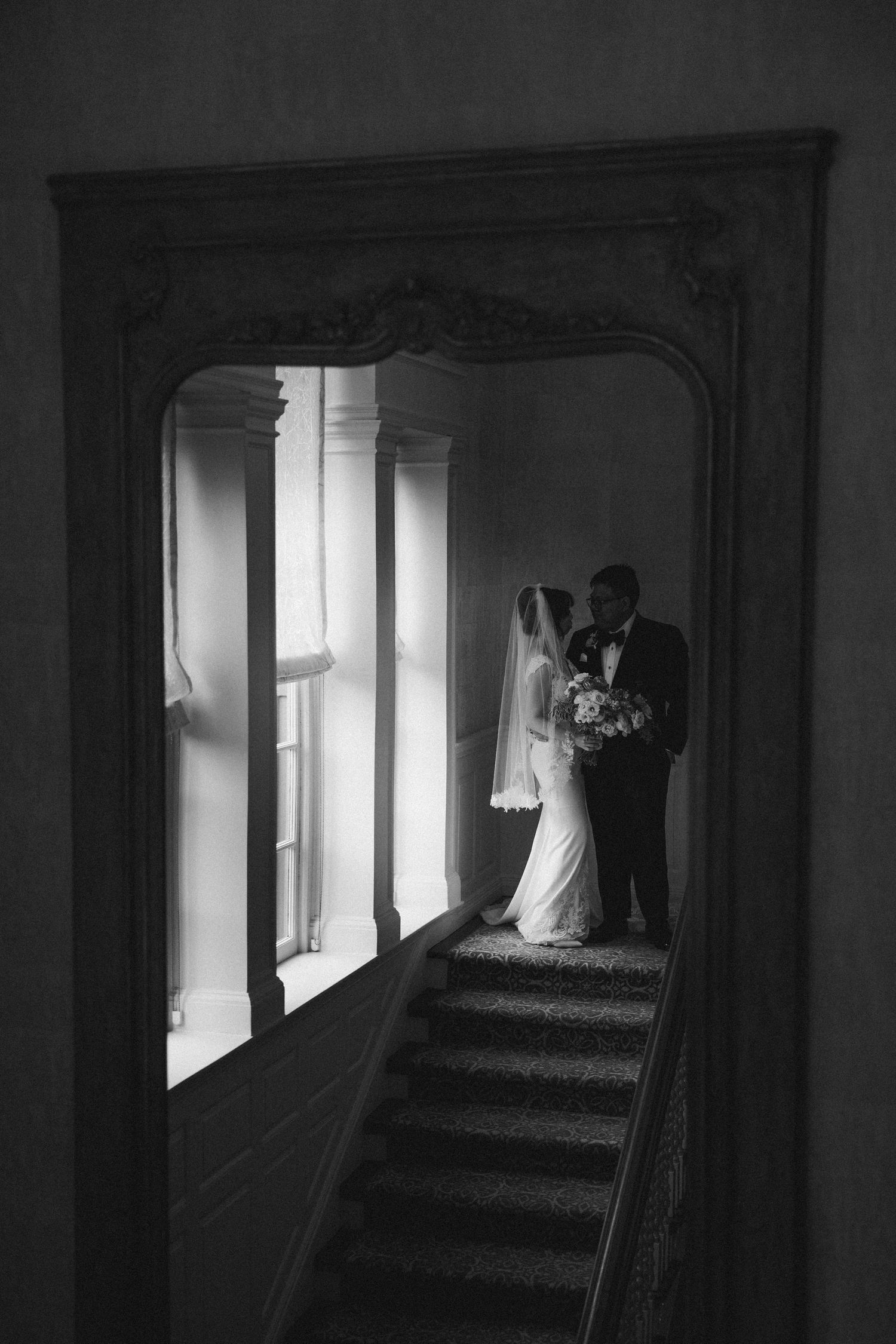 Silhouette of newlyweds embracing in black and white on elegant staircase framed by ornate archway and window light.