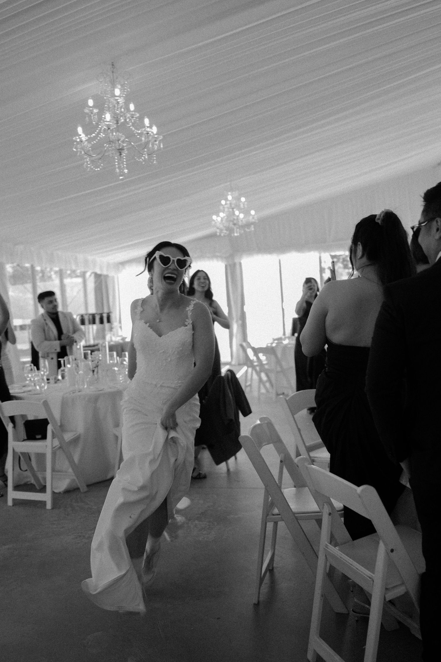 Black and white photo of a wedding reception inside an elegant tent with chandeliers and white chairs.
