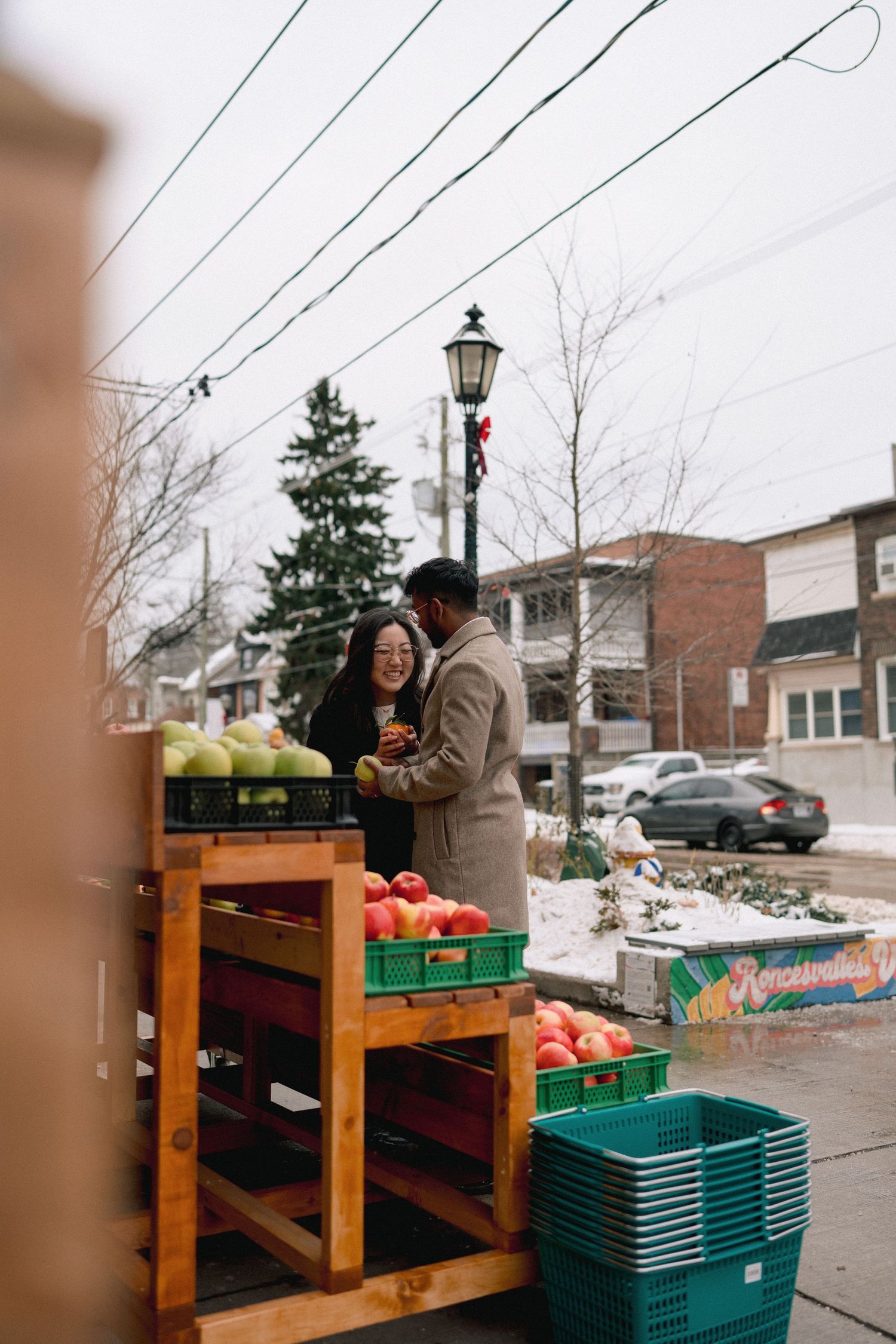 People gather at a wooden stand with green plastic bins and produce crates in a snowy winter setting.