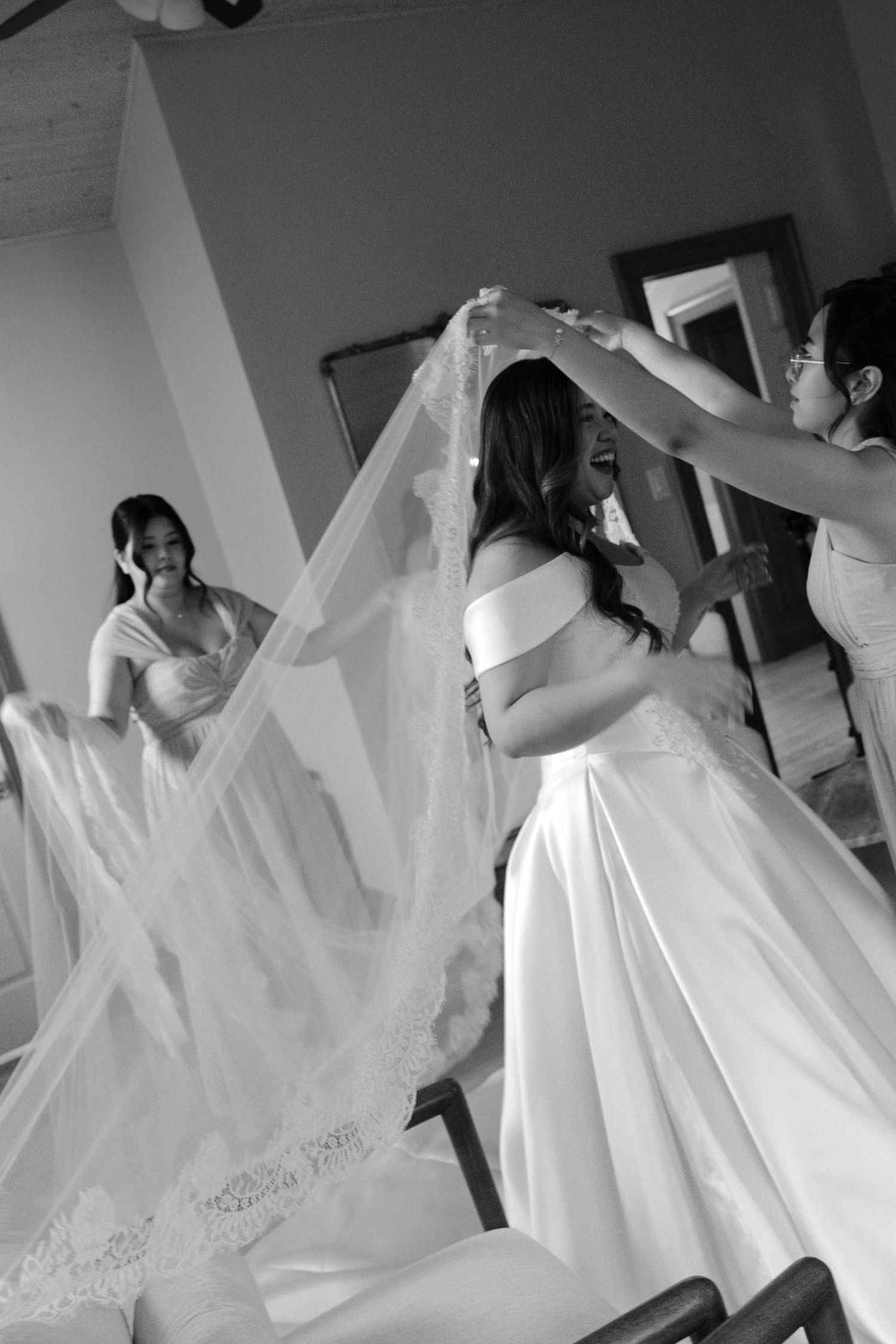 Black and white photo of bridesmaids helping a smiling bride put on her lace wedding veil before the ceremony.