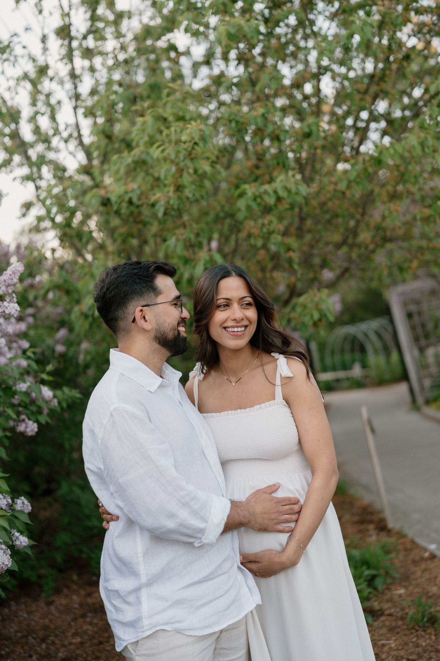 Smiling pregnant couple in white outfits embracing outdoors among lush green trees and purple lilac bushes.