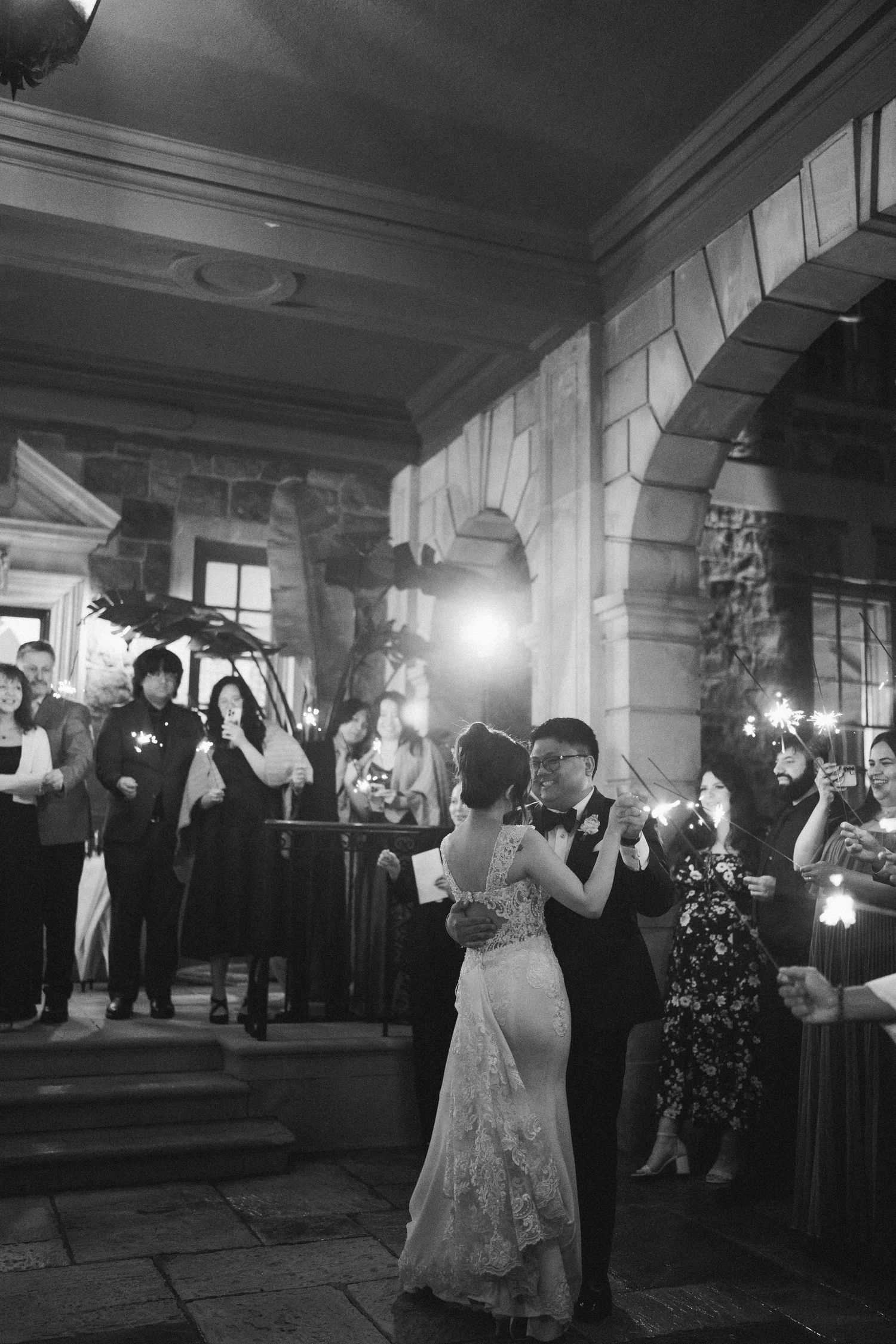 Black and white photo of a couple dancing their first dance at a wedding ceremony while guests watch with sparklers.