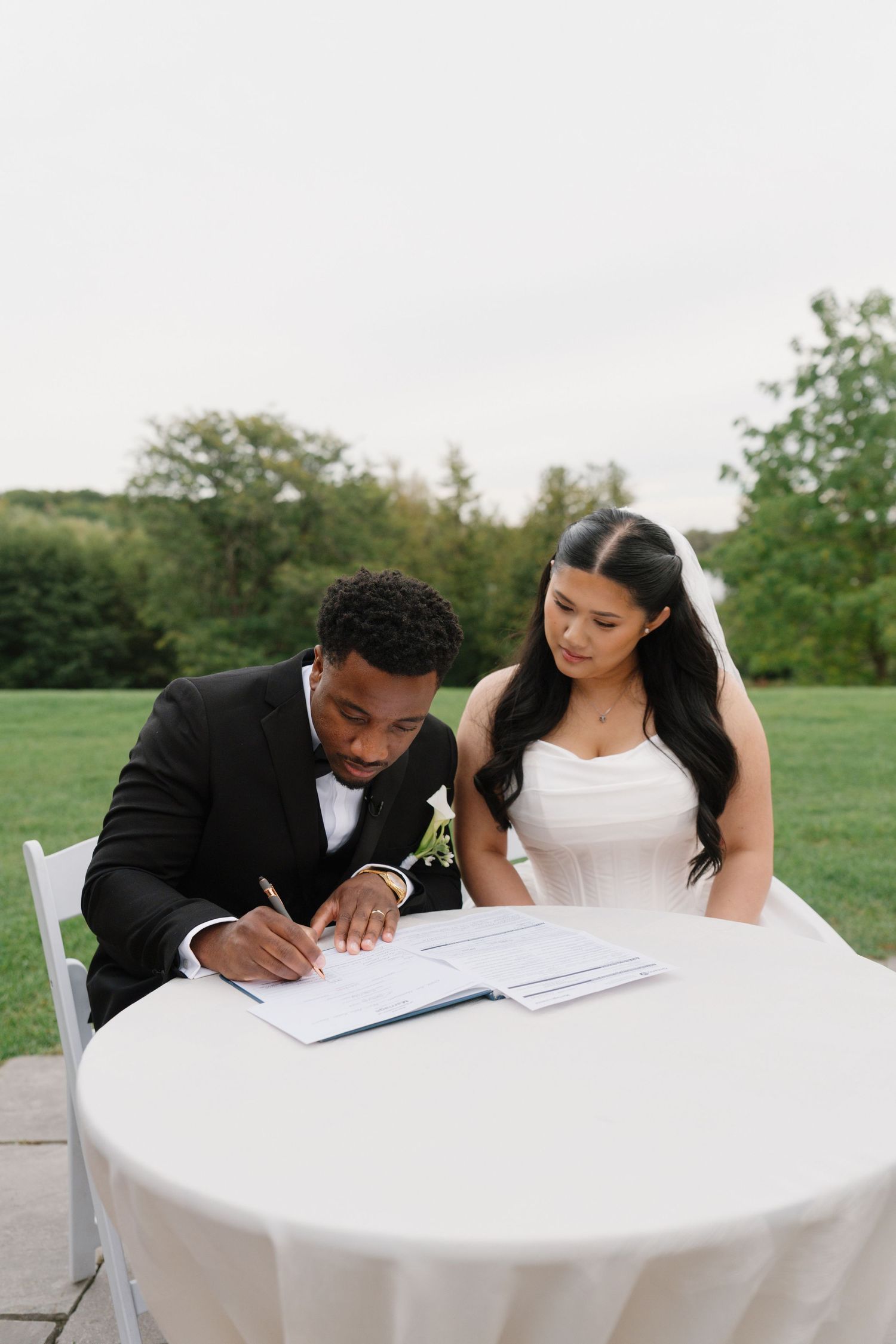 Bride and groom signing wedding certificate outdoors at white table, groom in black tuxedo, bride in white strapless gown with veil.