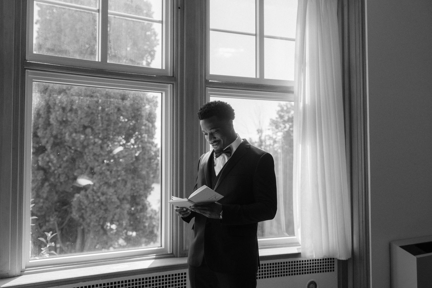 Groom in tuxedo reads notes by window in black and white wedding photo.