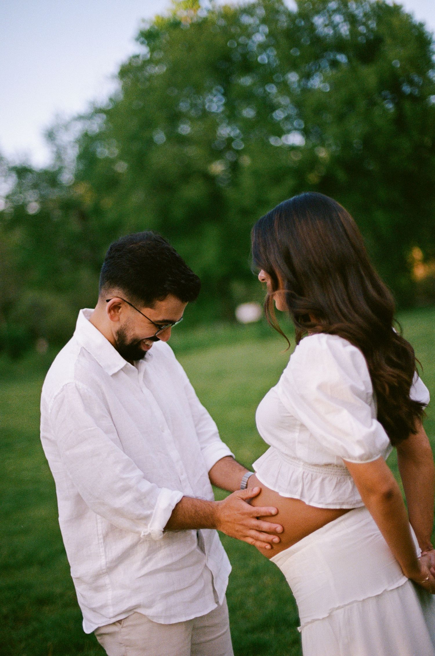 Partner tenderly touching pregnant womans bare belly outdoors in golden hour light during maternity session.