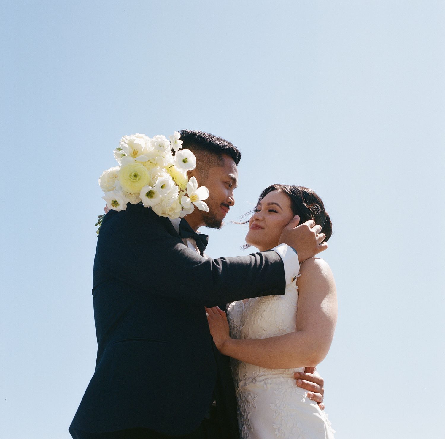 Newlywed couple sharing an intimate moment against blue sky at outdoor wedding ceremony.