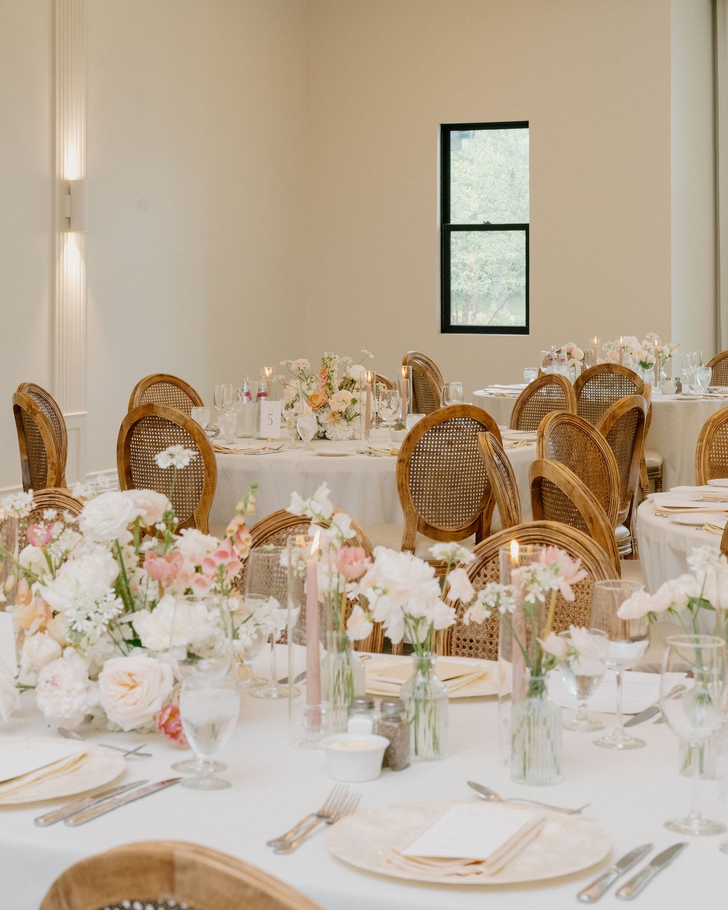 Elegant wedding reception with rattan chairs, white floral centerpieces, and soft pink blooms on white linen tables.