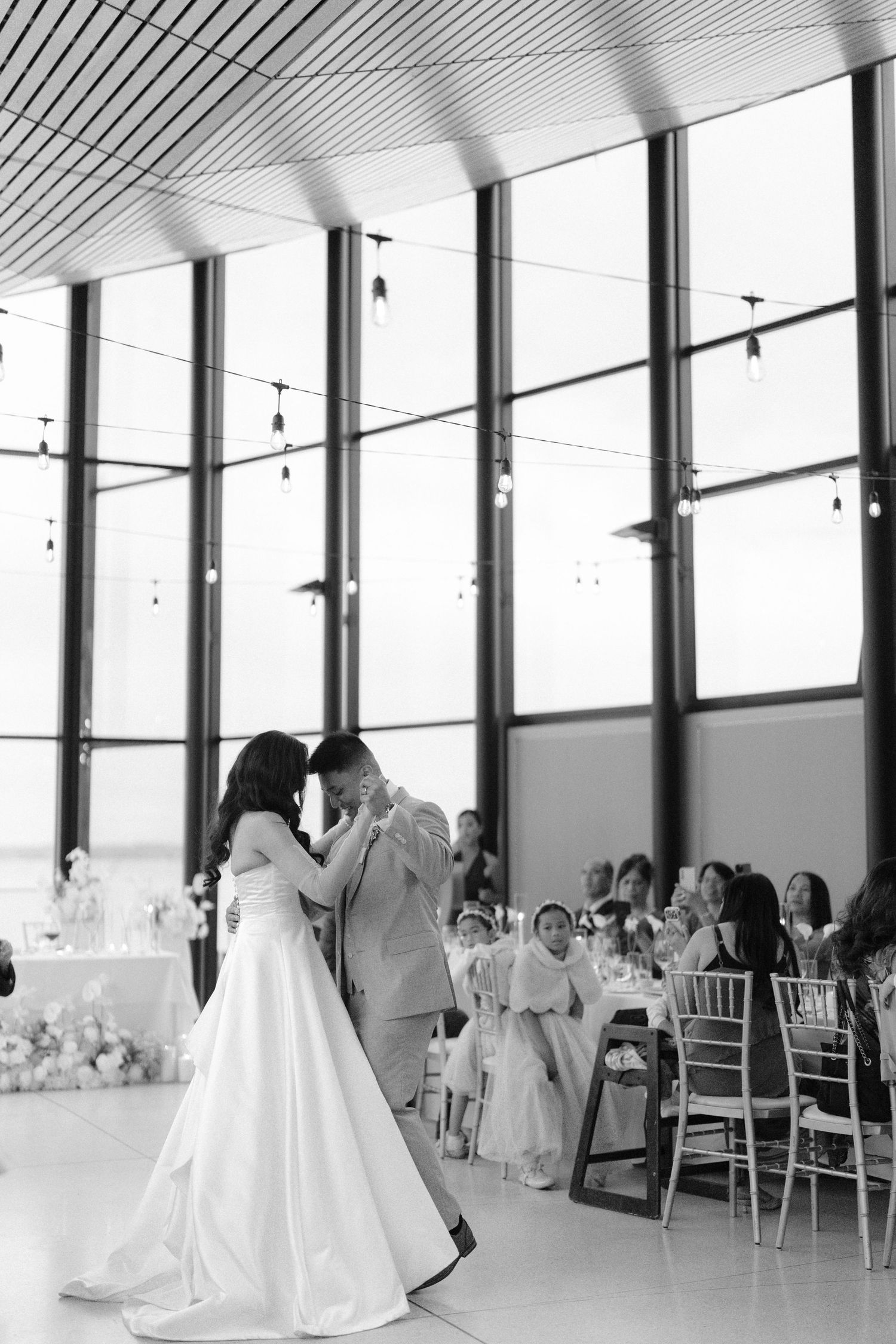 Black and white photo of bride and groom sharing first dance at elegant wedding reception with floor-to-ceiling windows.