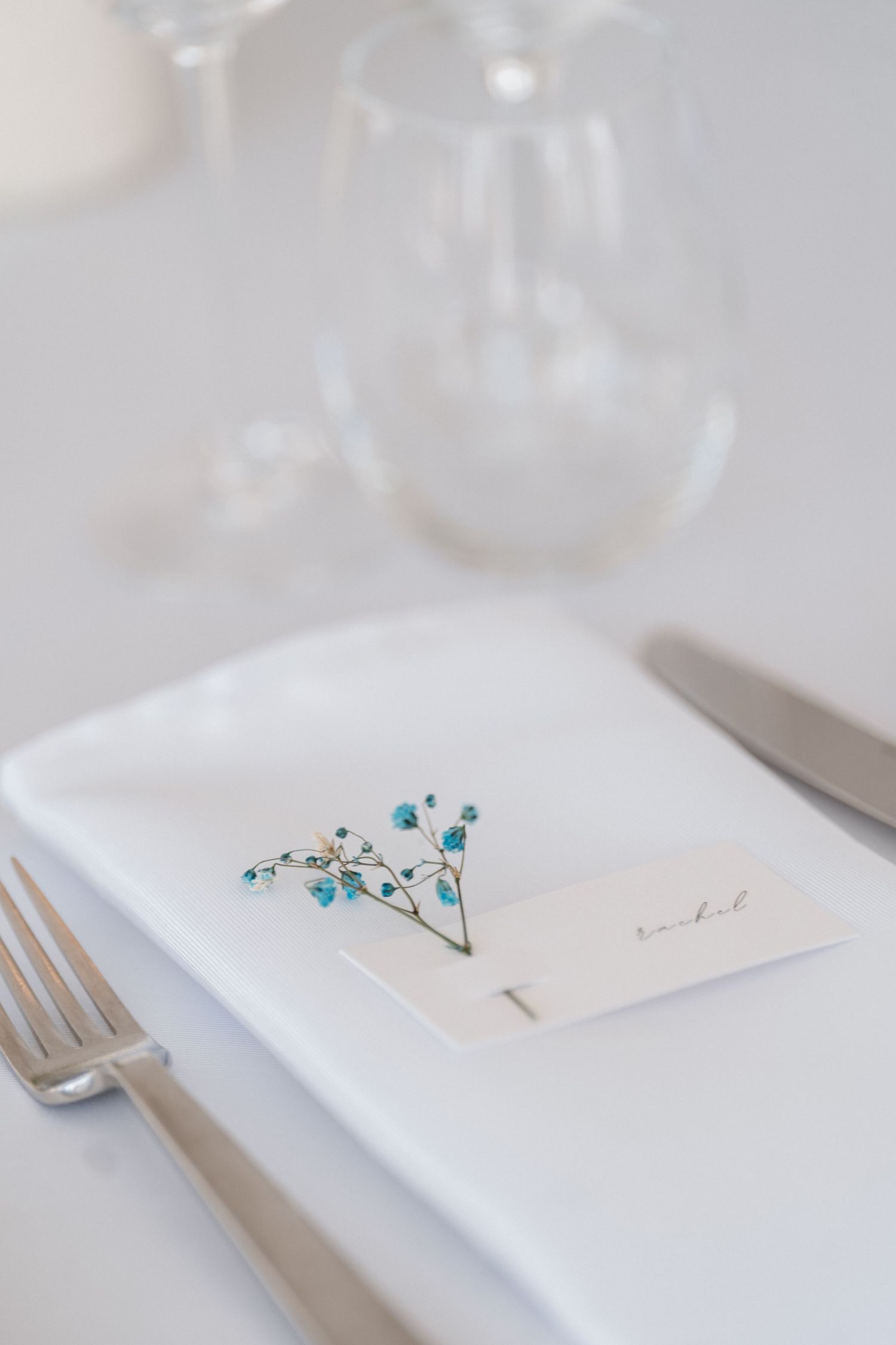 Elegant place setting with white napkin, silverware and small blue baby's breath flowers beside name card on table.
