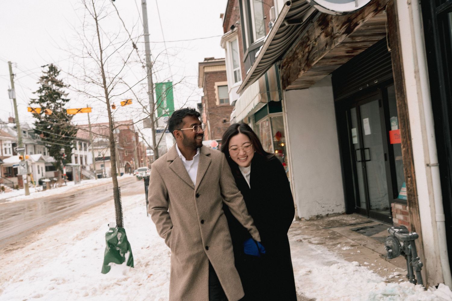 A couple bundled up in winter coats share a moment together on a snowy city sidewalk.
