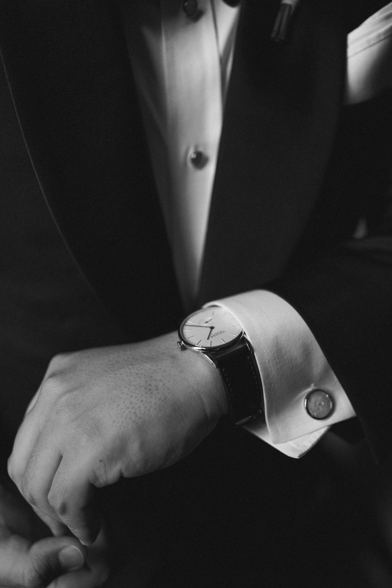 Black and white photo of a person wearing a formal suit checking an elegant wristwatch with white cuff and cufflink.
