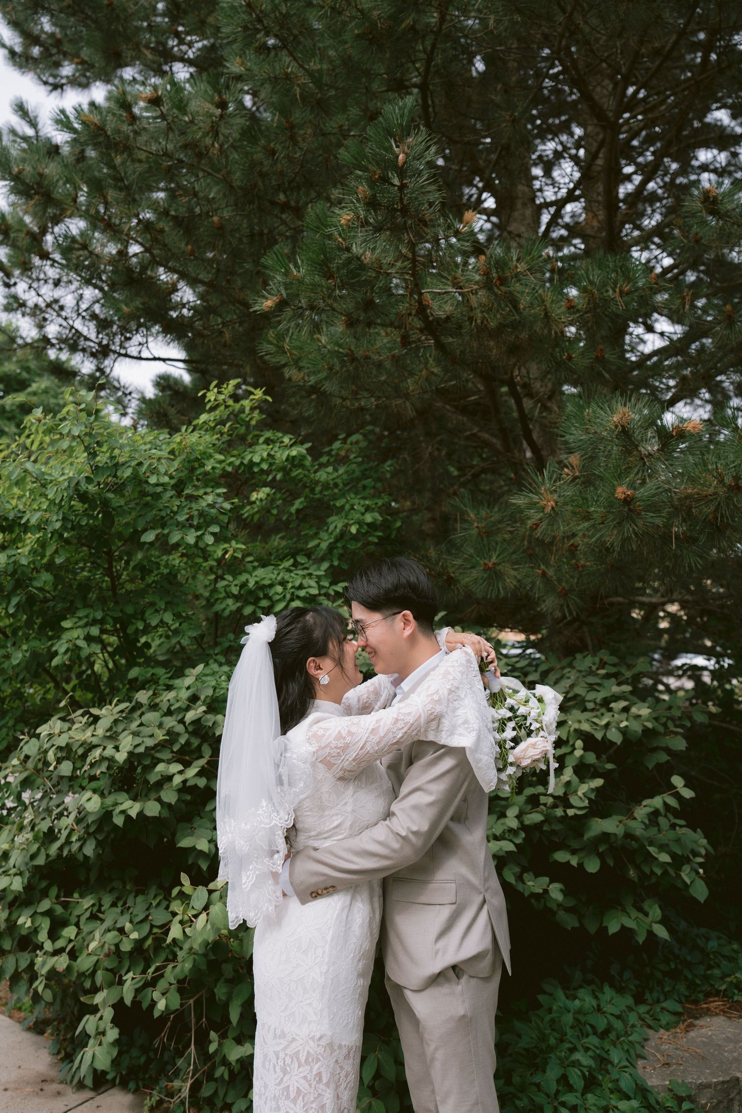 Bride and groom embracing outdoors in front of lush green trees, bride wearing lace gown and veil.