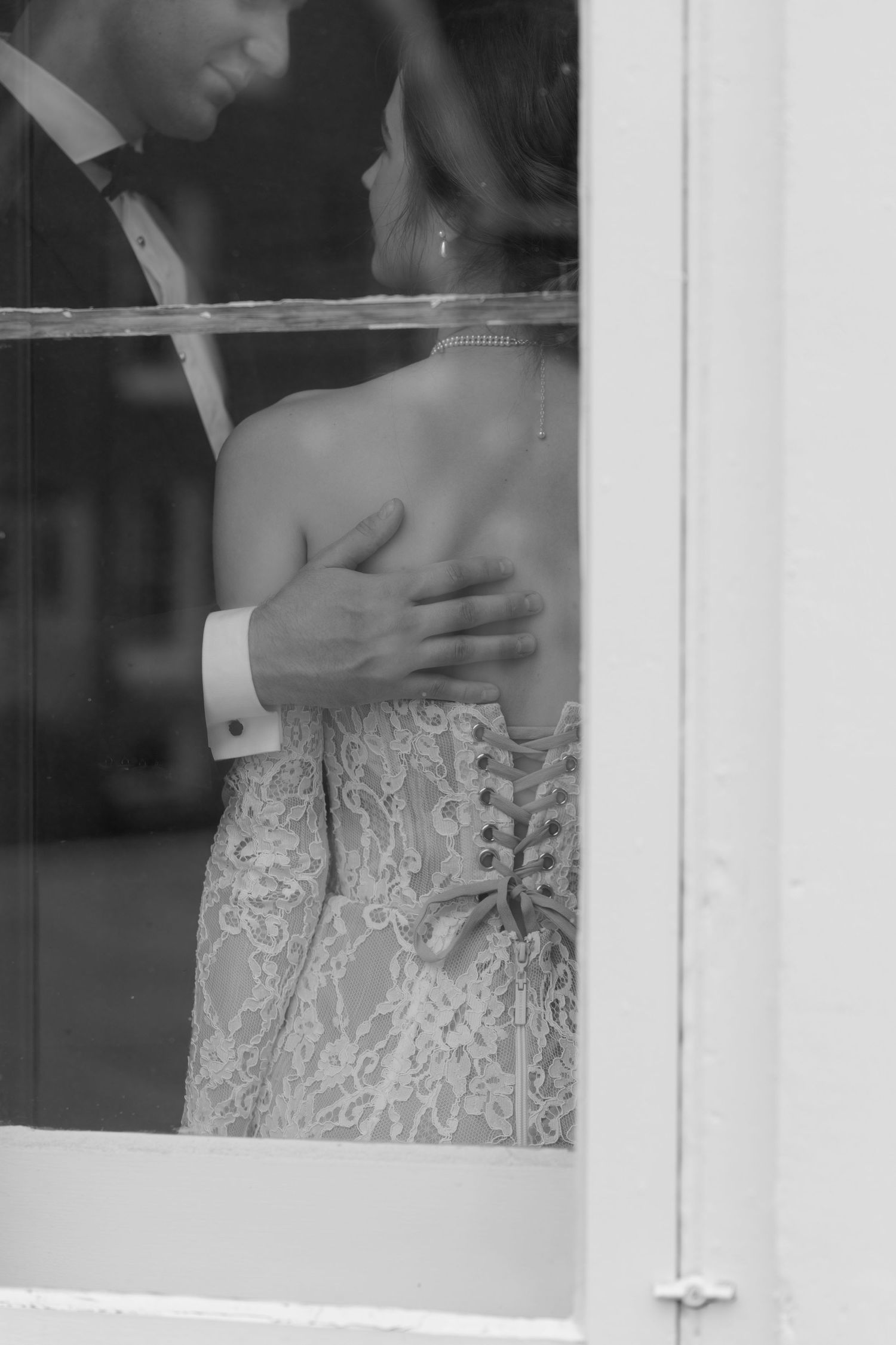 Black and white photo of bride and groom embracing behind a window, showing lace dress detail.