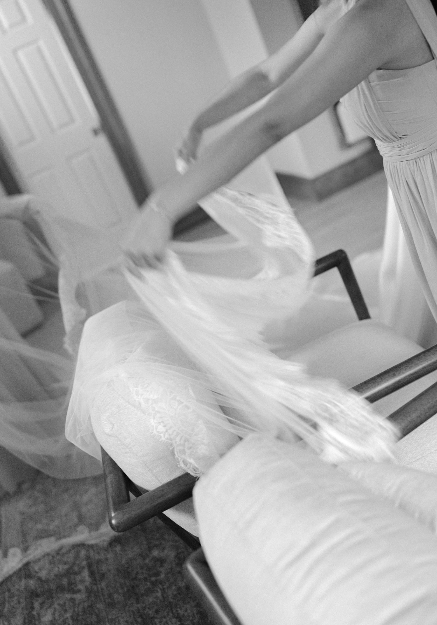 Black and white photo of a bride arranging her flowing white wedding veil and gown on a chair in a bridal suite.