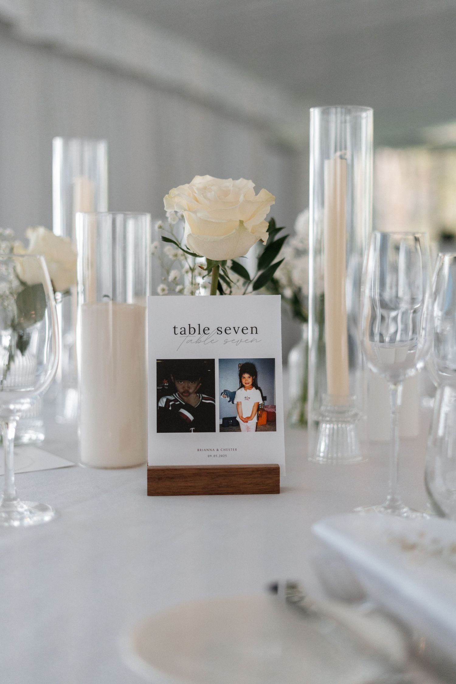 Wedding table centerpiece with photo frame table number, white rose, and glass candle holders on crisp white tablecloth.