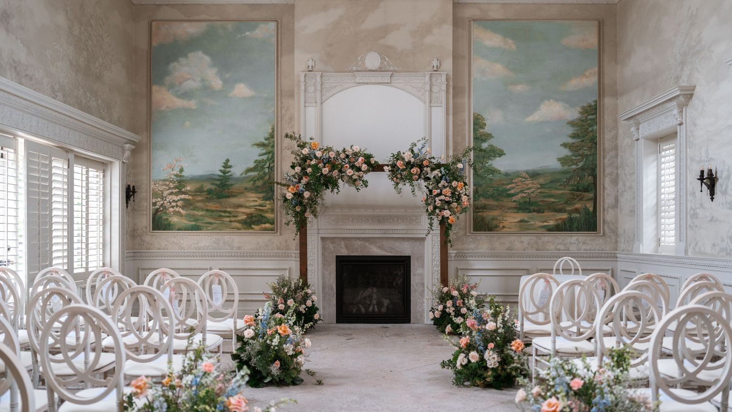 Elegant wedding ceremony setup with white chairs and floral arrangements around a fireplace between two landscape paintings.