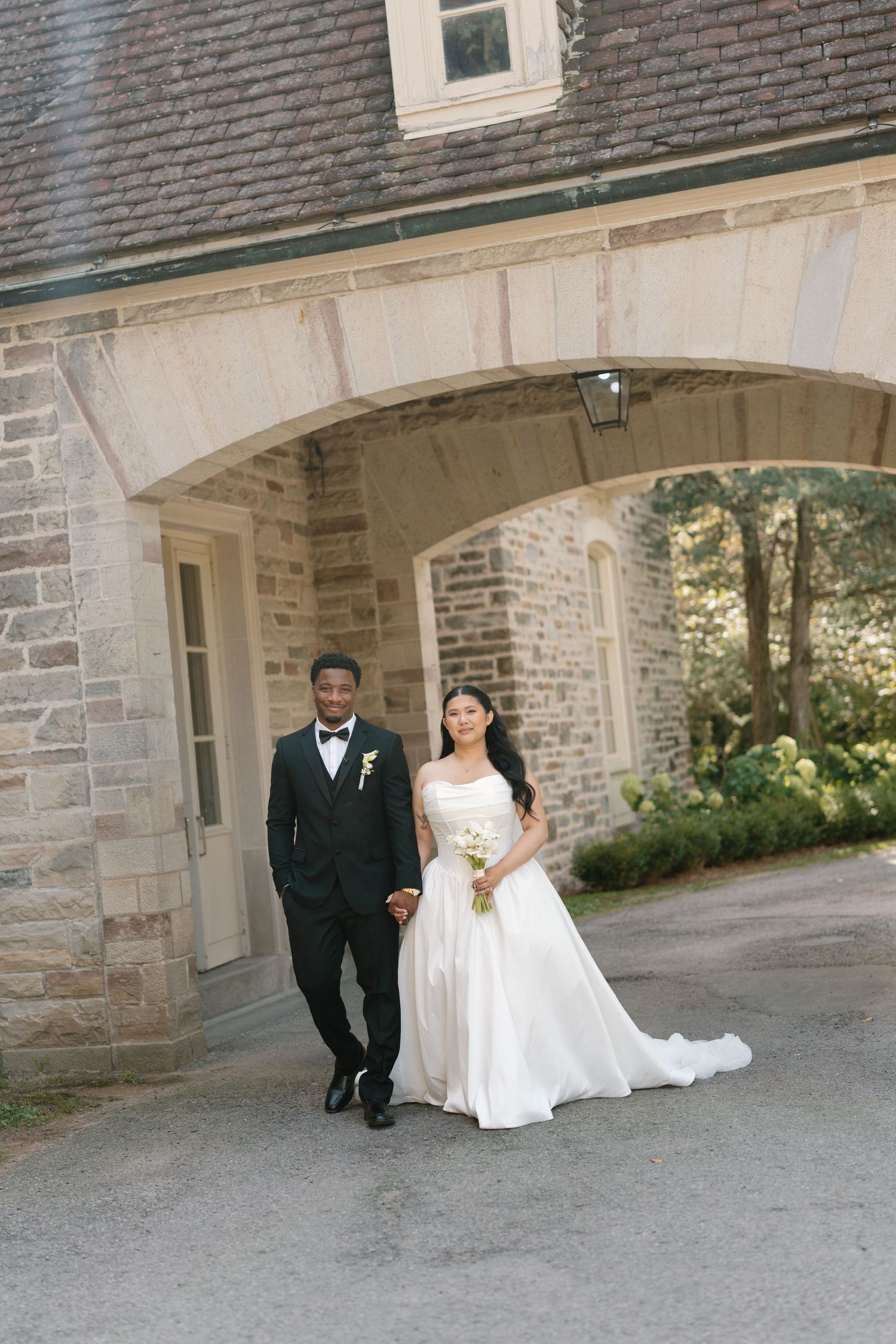 Bride in white gown and groom in tuxedo walk hand in hand under stone archway at elegant wedding venue.