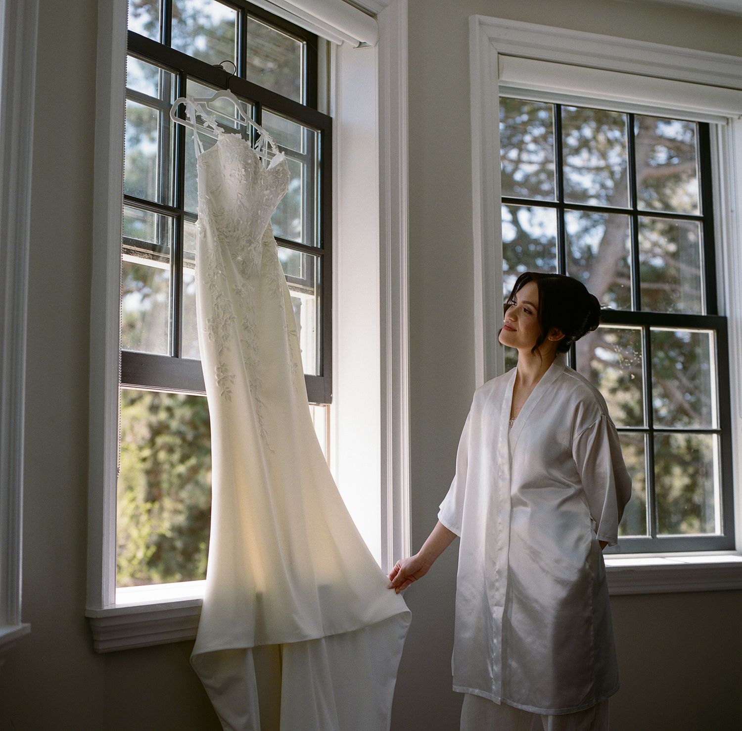 A white lace wedding dress hangs by a window while someone in a white robe admires it in natural light.