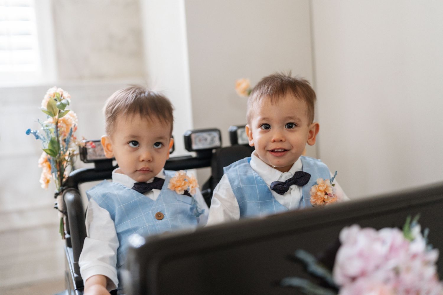 Two children in light blue vests and bow ties sit together at a piano with flowers nearby.