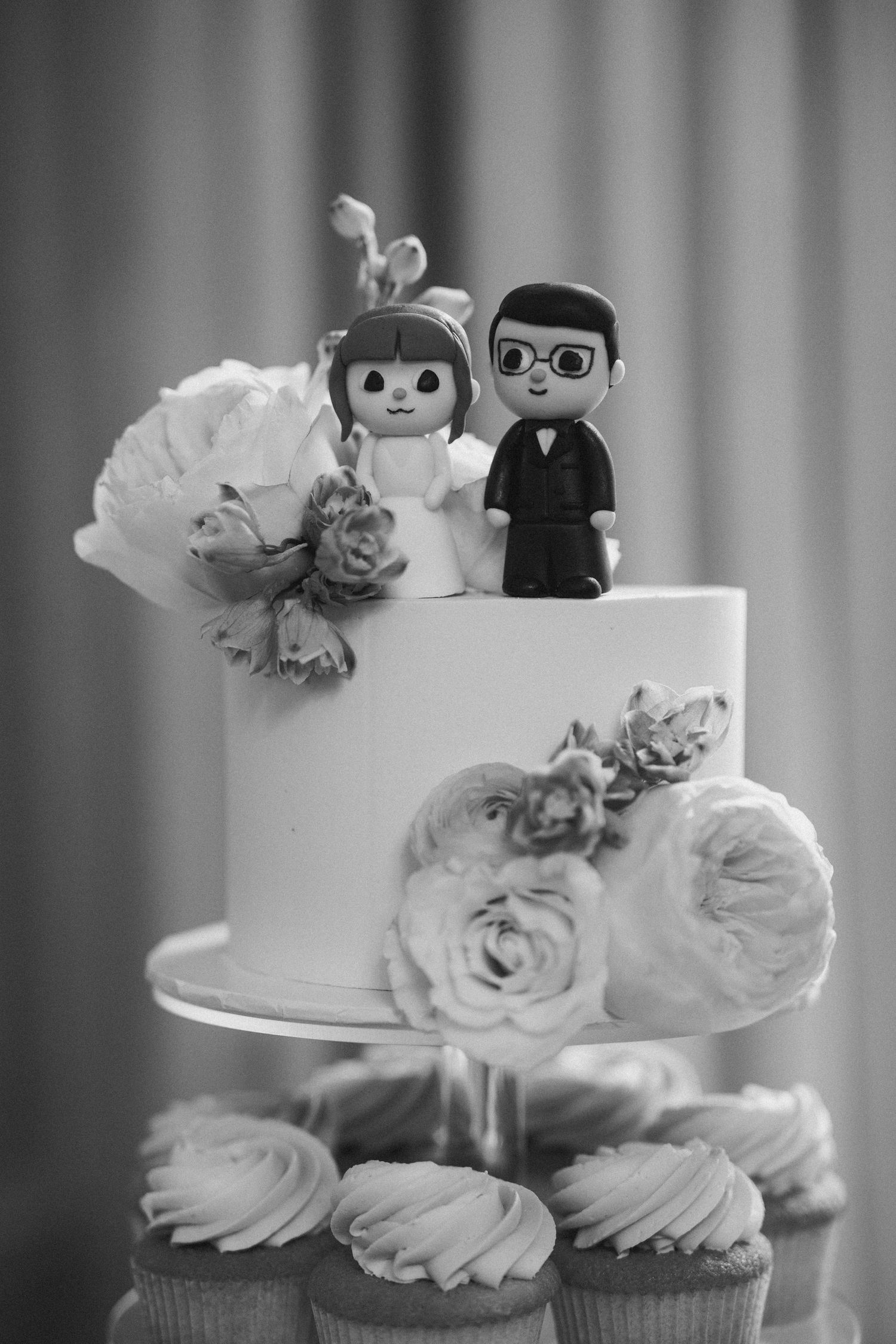 Black and white wedding cake topped with cartoon bride and groom figurines and decorated with roses.