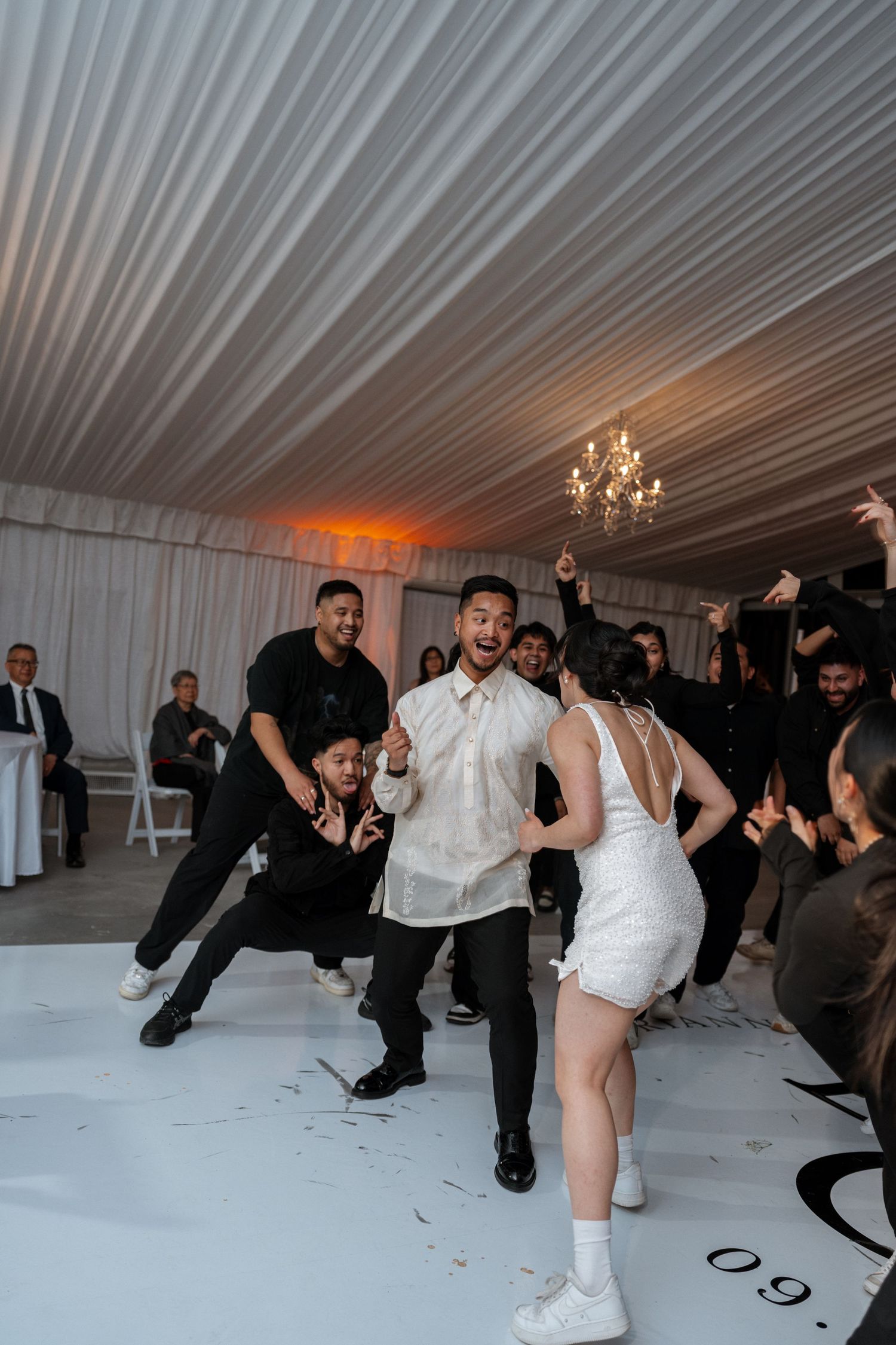 Wedding guests dance and celebrate on a white dance floor under a draped tent ceiling with chandelier lighting.