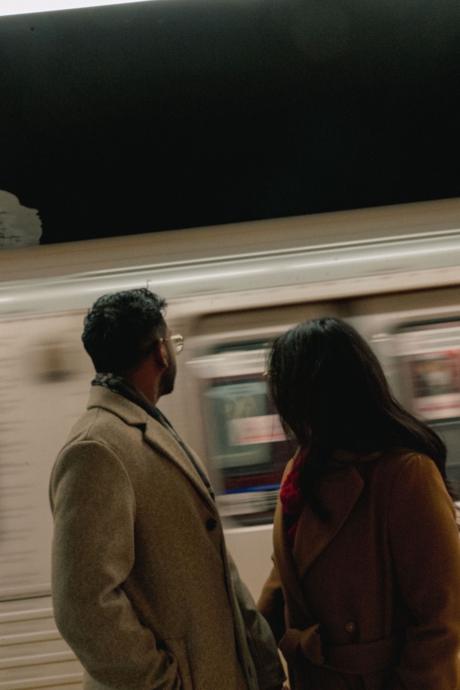Motion-blurred subway train passes by as two people wait on the platform in dark lighting conditions.