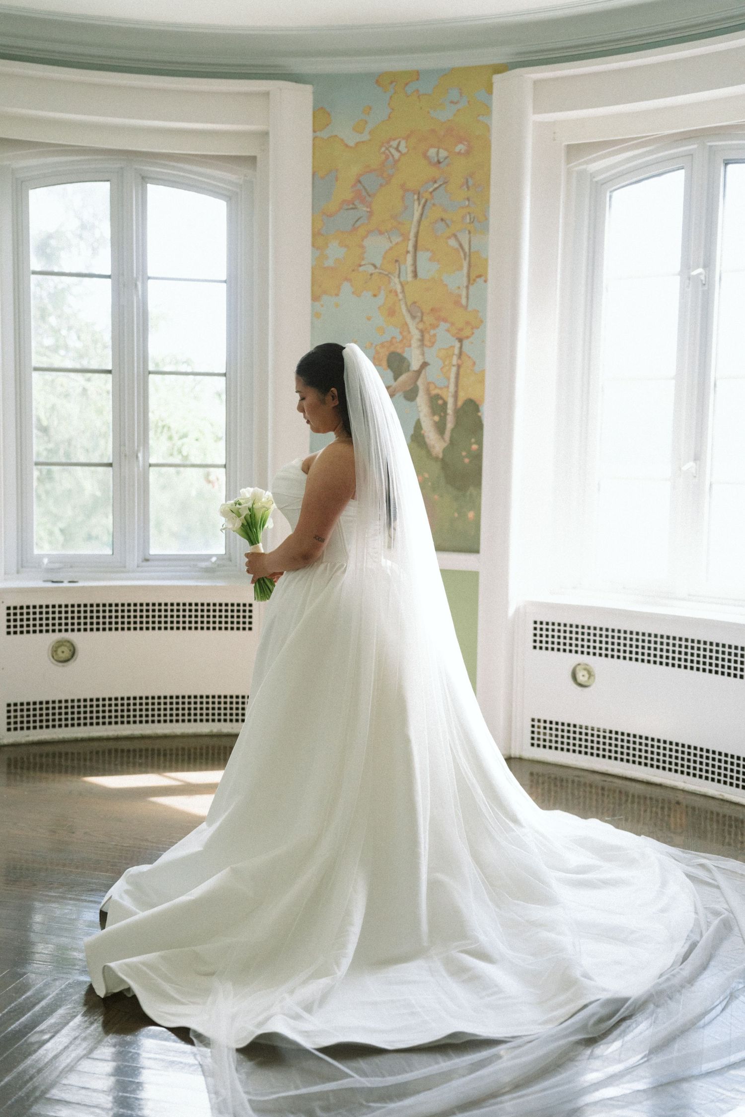 Bride in white gown and veil holding bouquet stands by sunlit windows with floral mural in elegant venue.