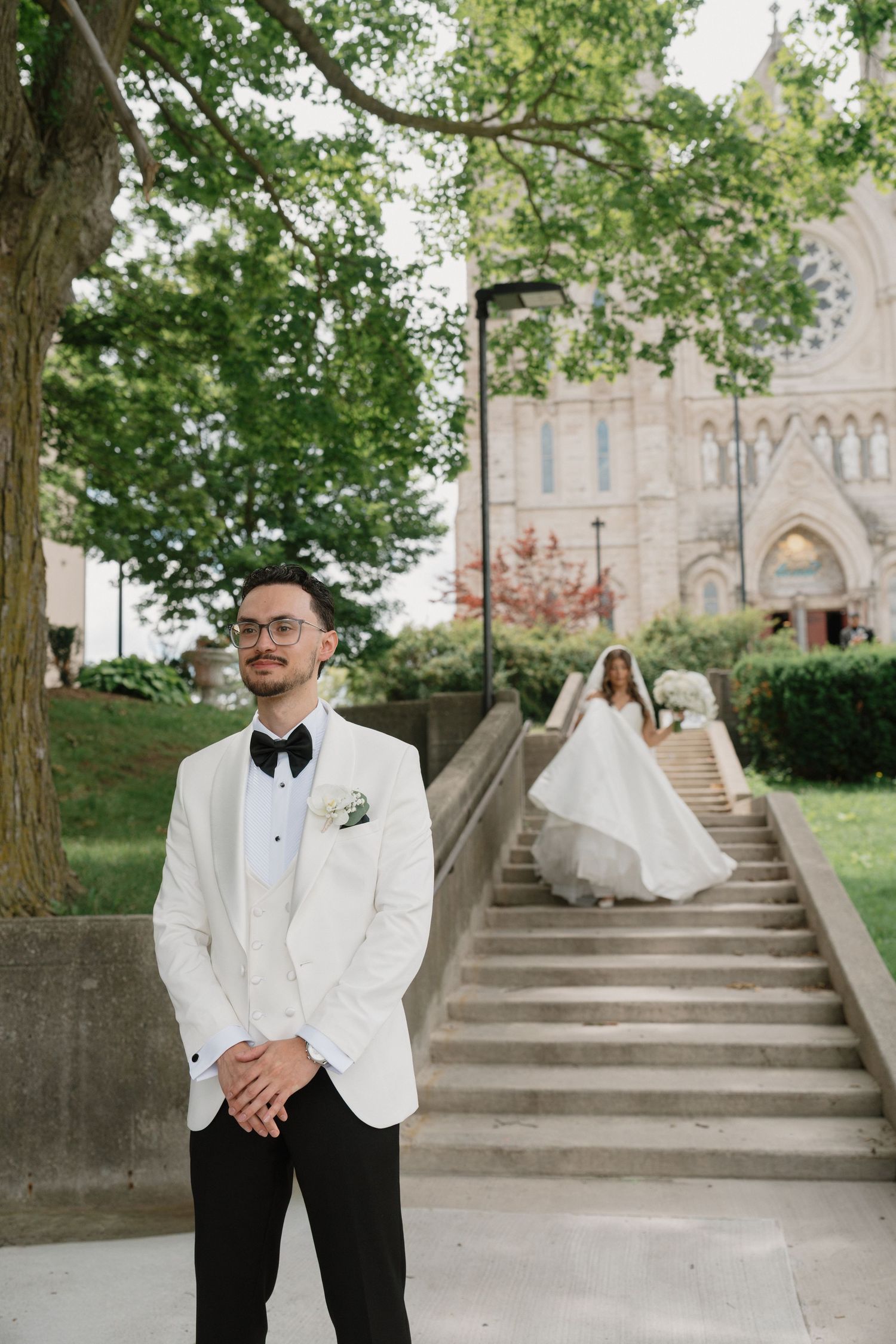 Groom in white tuxedo jacket stands outdoors as bride descends stone steps near a Gothic church.