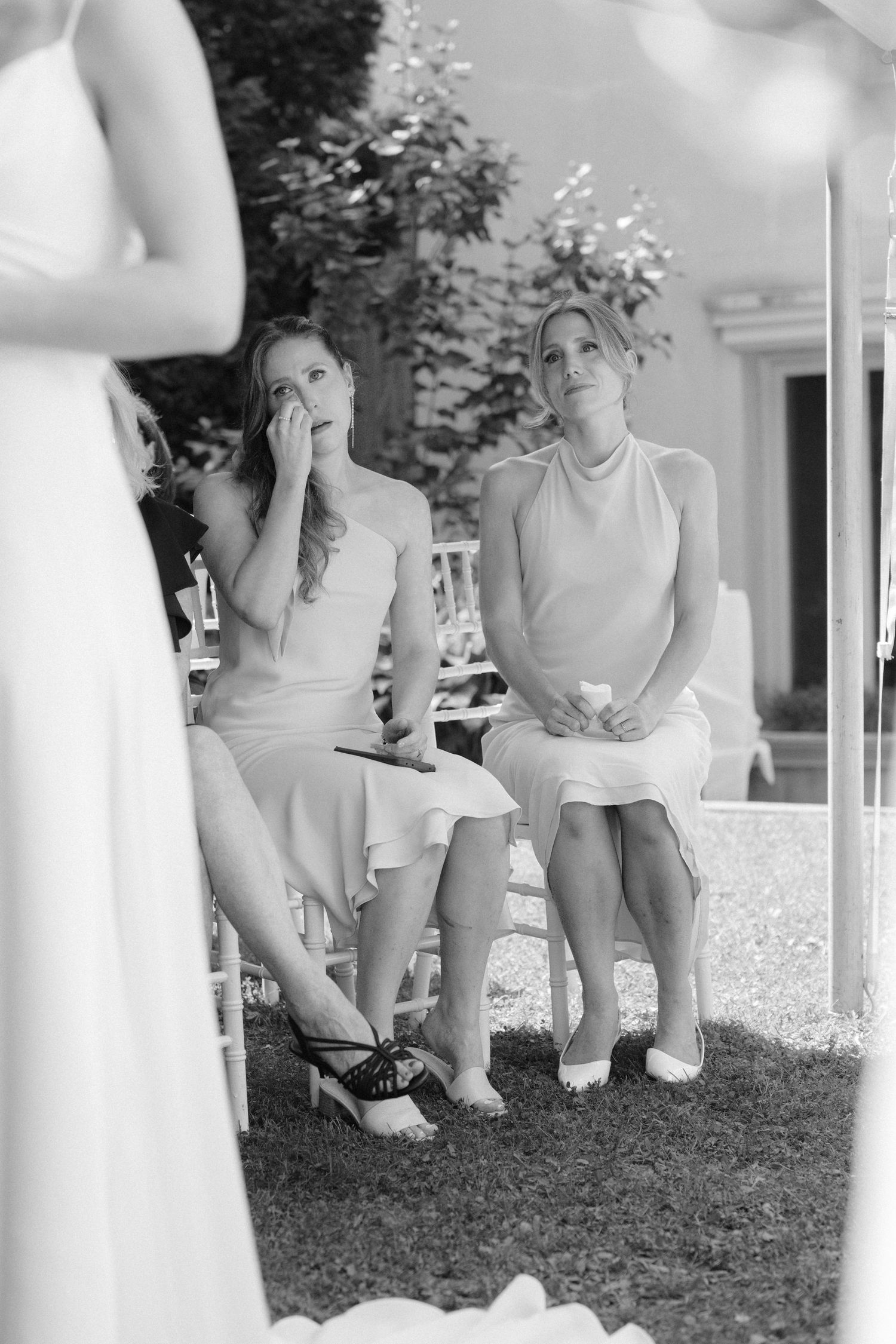 Black and white photo of two emotional female wedding guests seated outdoors, wiping tears during a ceremony.