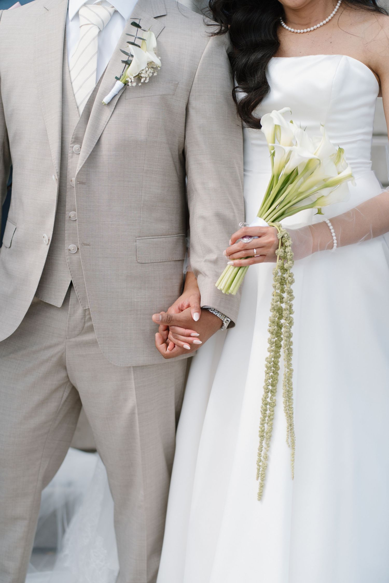 Bride and groom holding hands, she holds white calla lily bouquet, he wears beige suit with boutonniere.