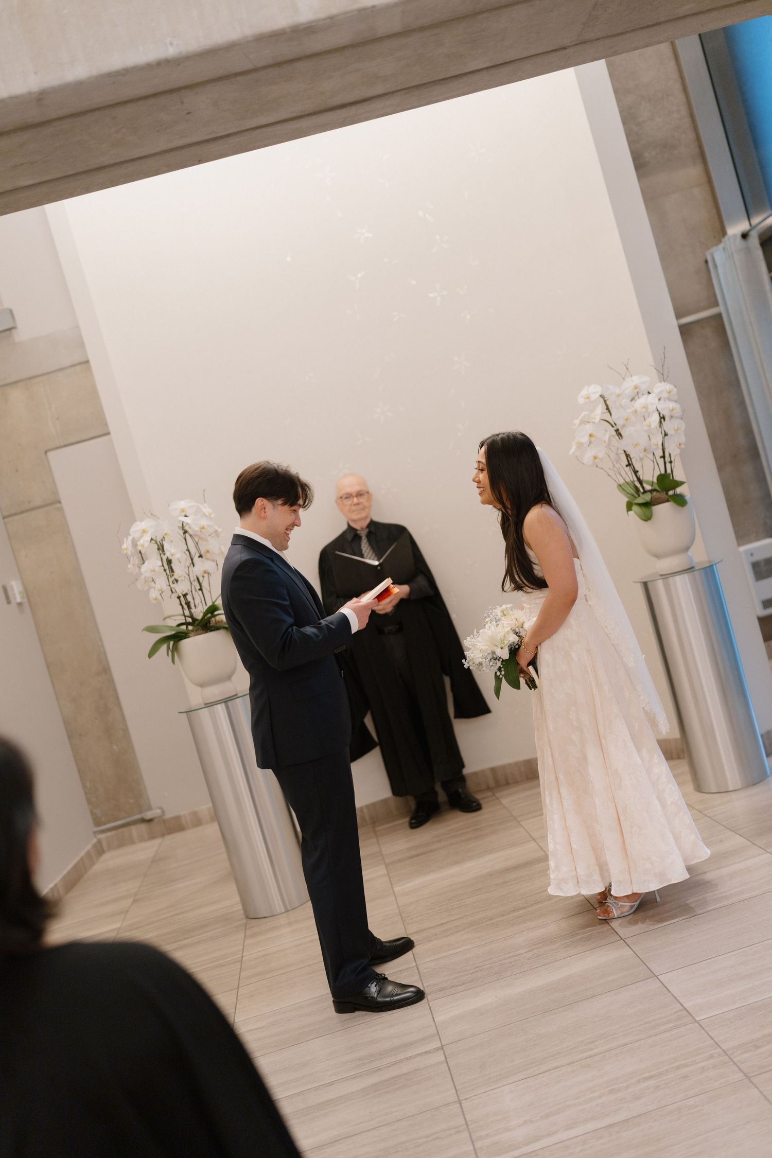 Couple exchanging vows at indoor wedding ceremony with officiant, white orchids, bride in white gown holding bouquet.
