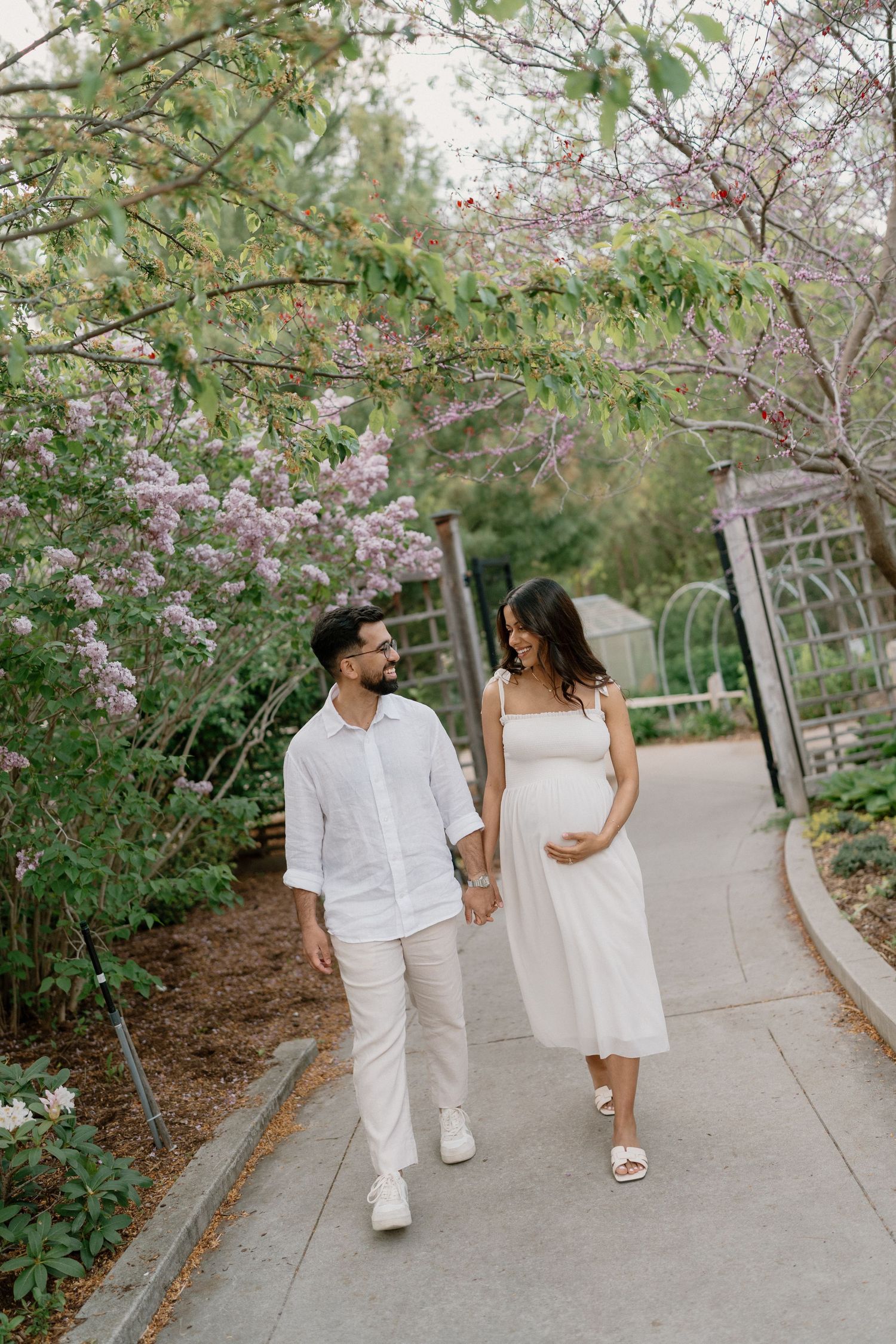 Smiling expectant couple walking hand in hand through a blooming garden with purple lilac flowers.