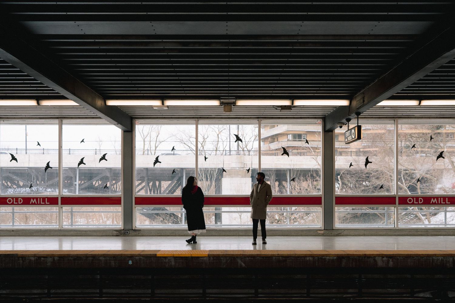 Two silhouettes stand on a train platform as birds fly past red and white wall art in the background.