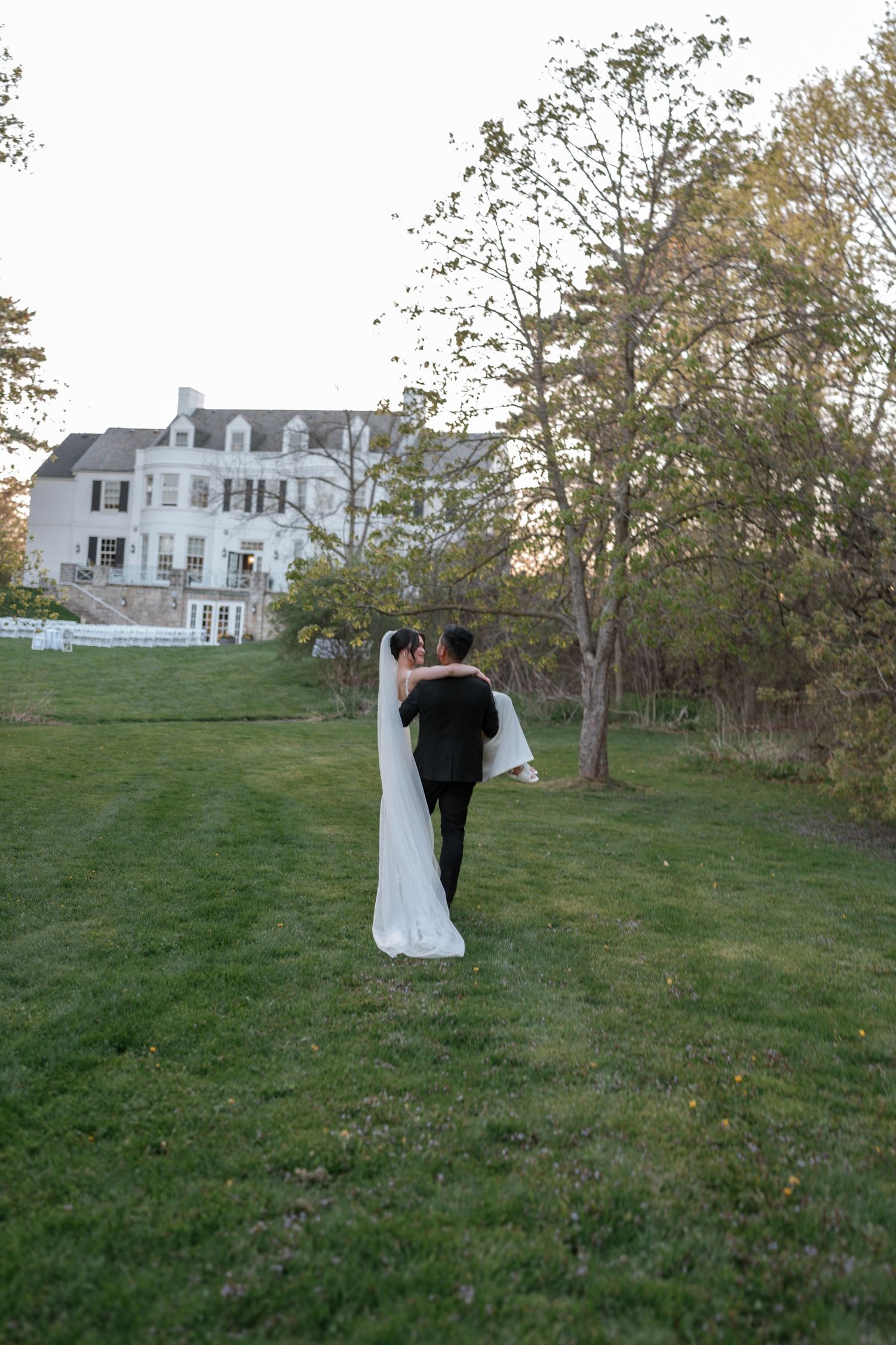 A couple embraces on the lawn of an elegant white mansion during an autumn evening wedding celebration.