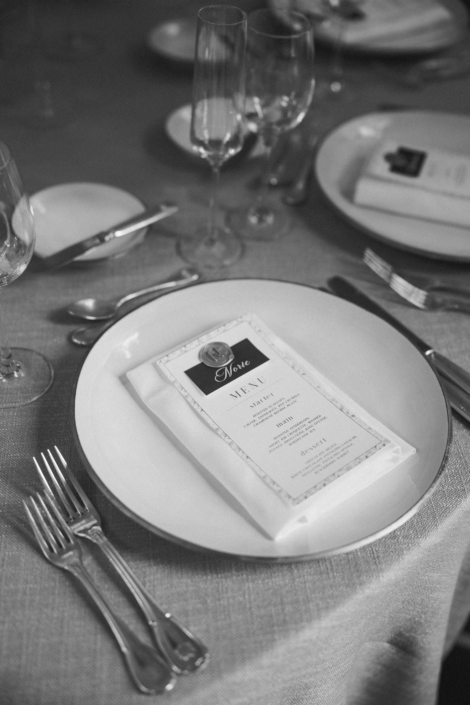 Elegant black and white table setting with menu card, white plate, wine glass and formal silverware on linen tablecloth.