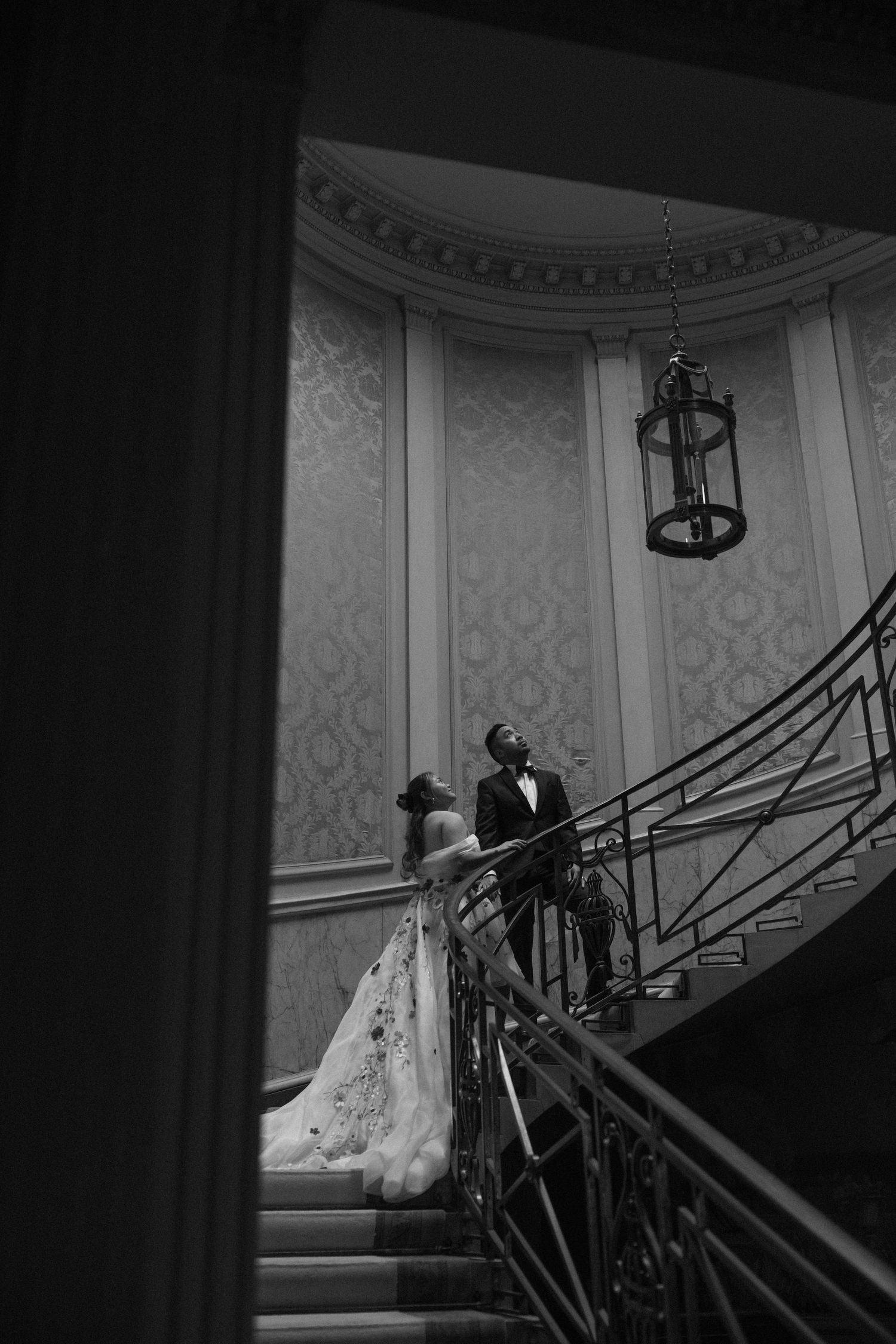 Black and white photo of bride and groom on grand staircase in ornate venue.