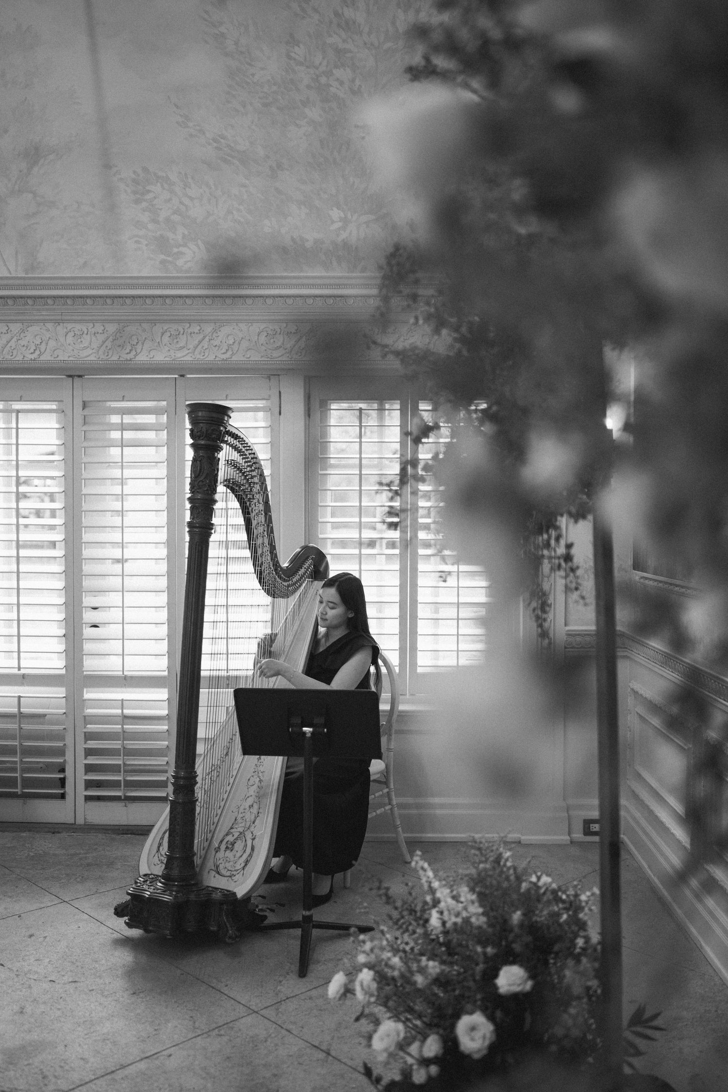 A harpist performs in an elegant interior setting with white shuttered windows and floral decor in black and white tones.