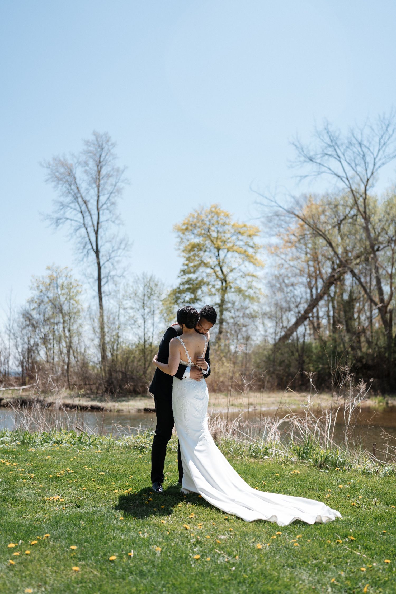 A couple in wedding attire embraces on a grassy lawn beside a pond with bare trees in the background on a sunny day.
