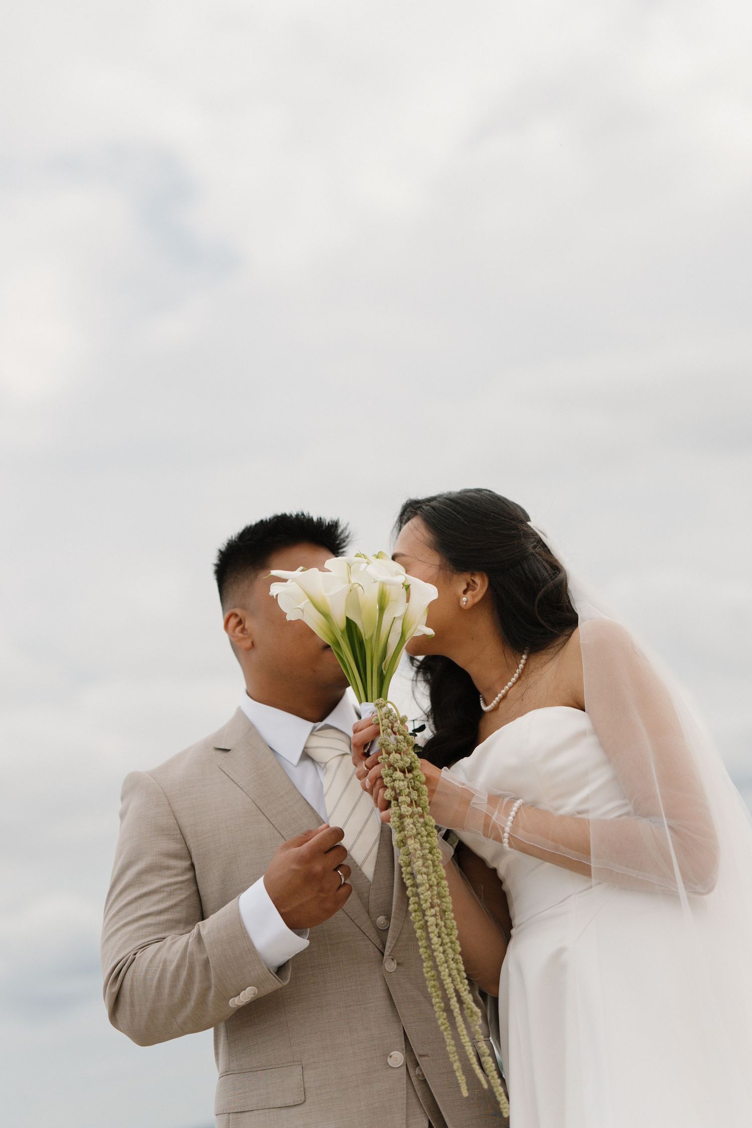 A bride and groom share a romantic moment, kissing behind a white calla lily bouquet outdoors on their wedding day.