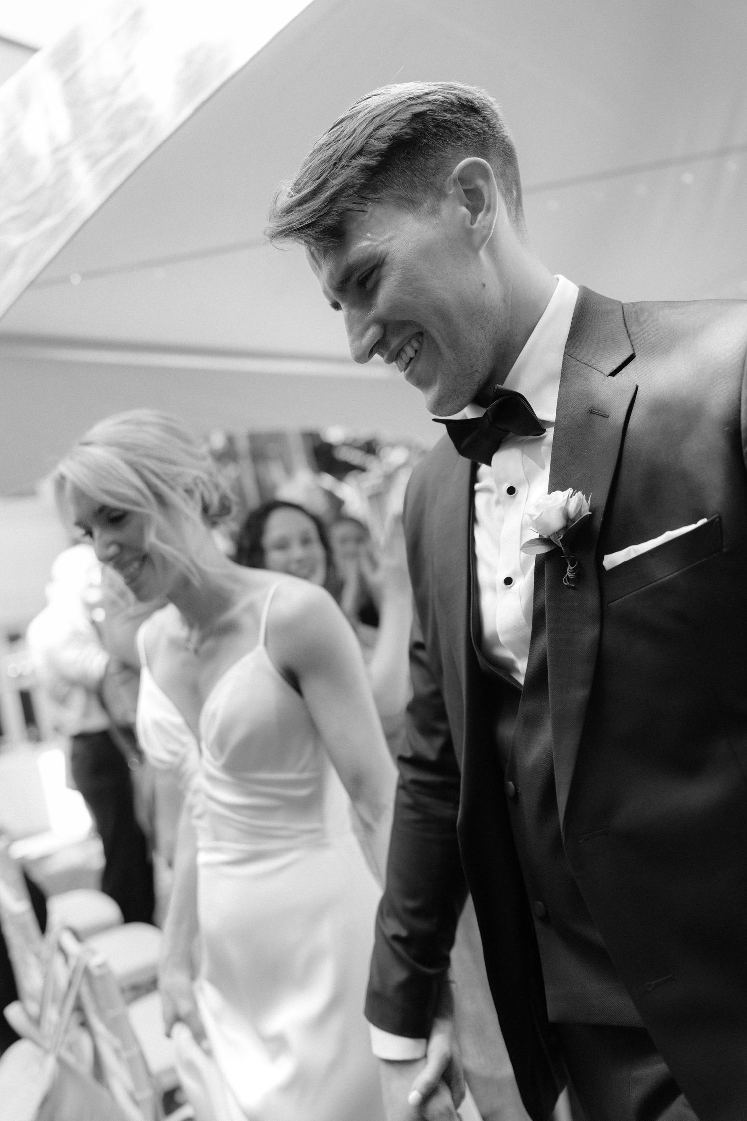 Black and white photo of a smiling groom in a tuxedo walking with his bride in a white dress at their wedding.