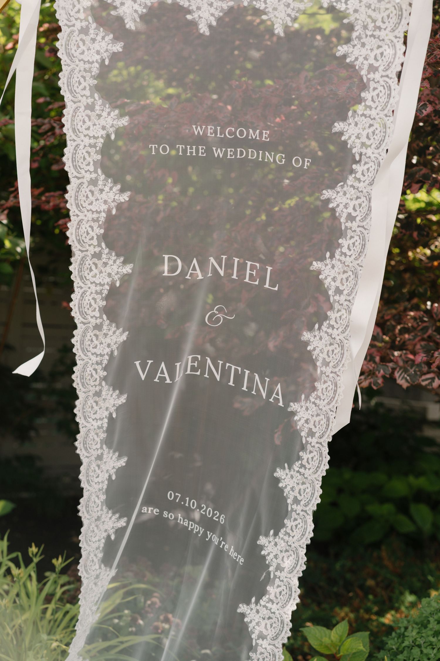 Elegant lace wedding welcome sign for Daniel and Valentina, dated 07.10.2026, displayed outdoors against greenery.