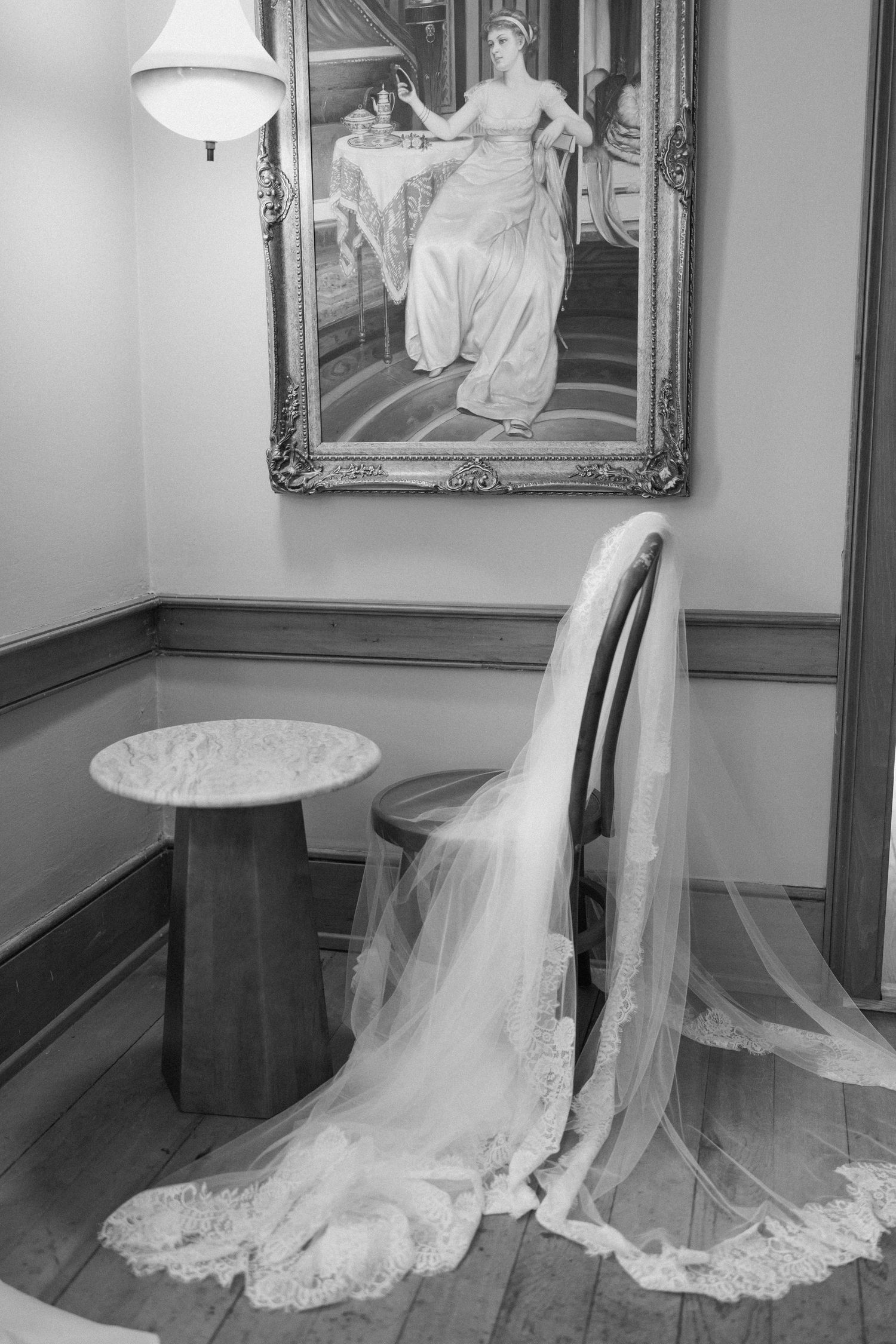 Black and white photo of a bridal veil draped over a chair beside a painting of a woman in a gown.