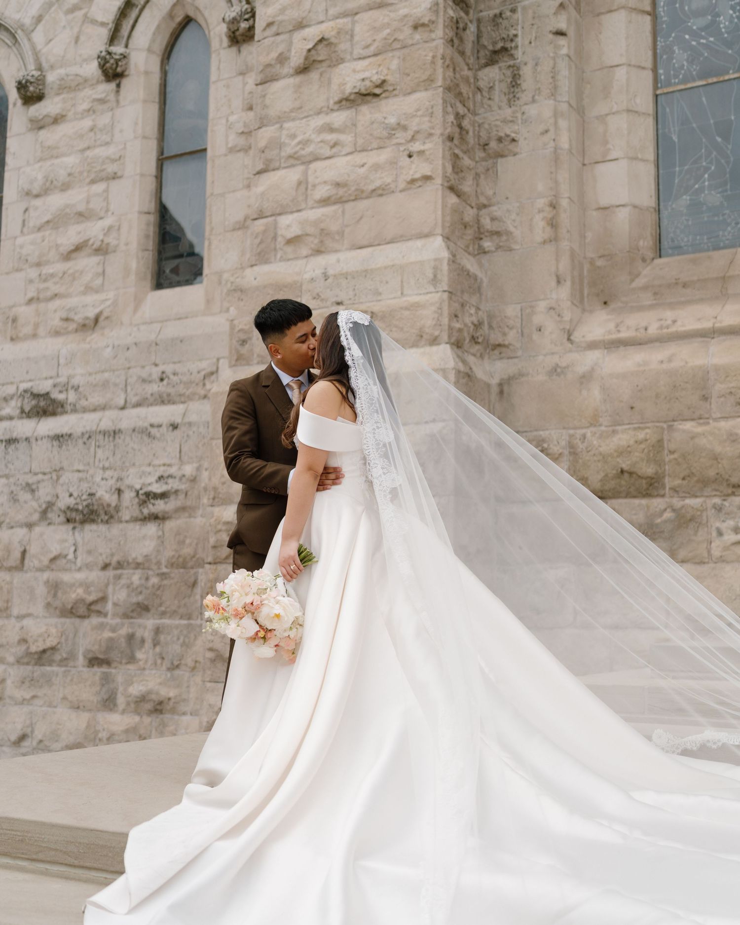 Bride and groom share a romantic kiss on church steps, her elegant white gown and long veil trailing beautifully.