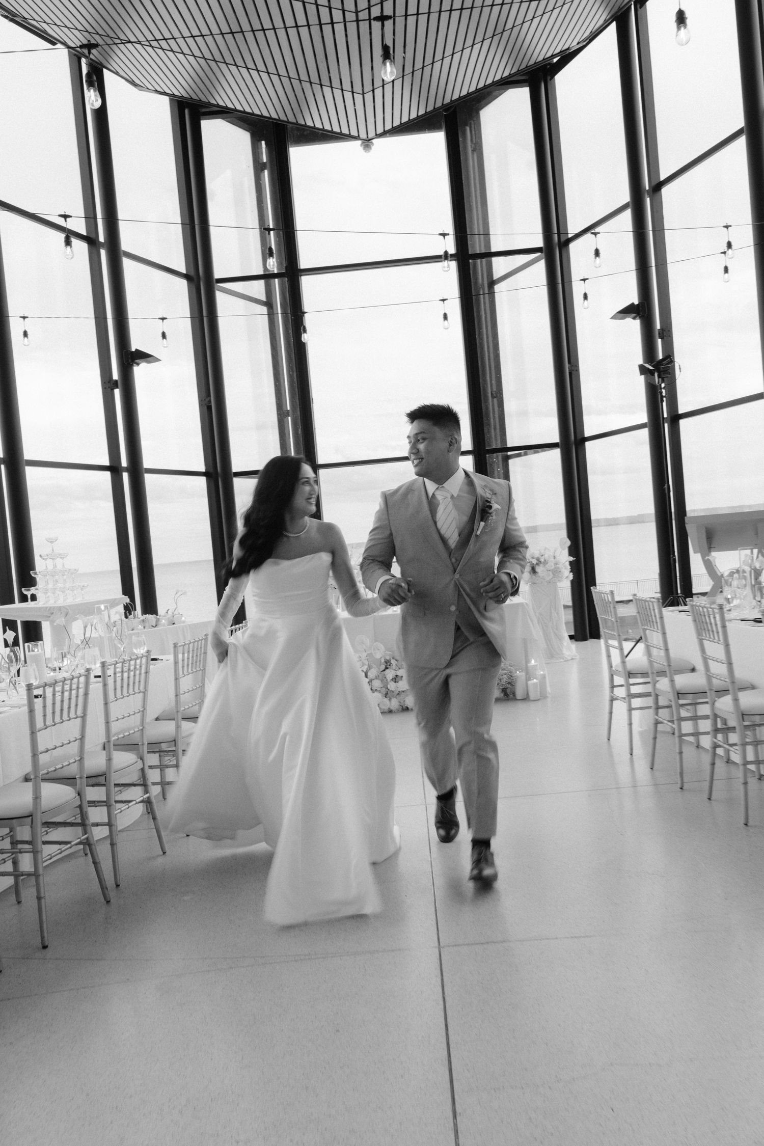 Black and white photo of a bride and groom walking hand in hand through an elegant glass-walled wedding venue.